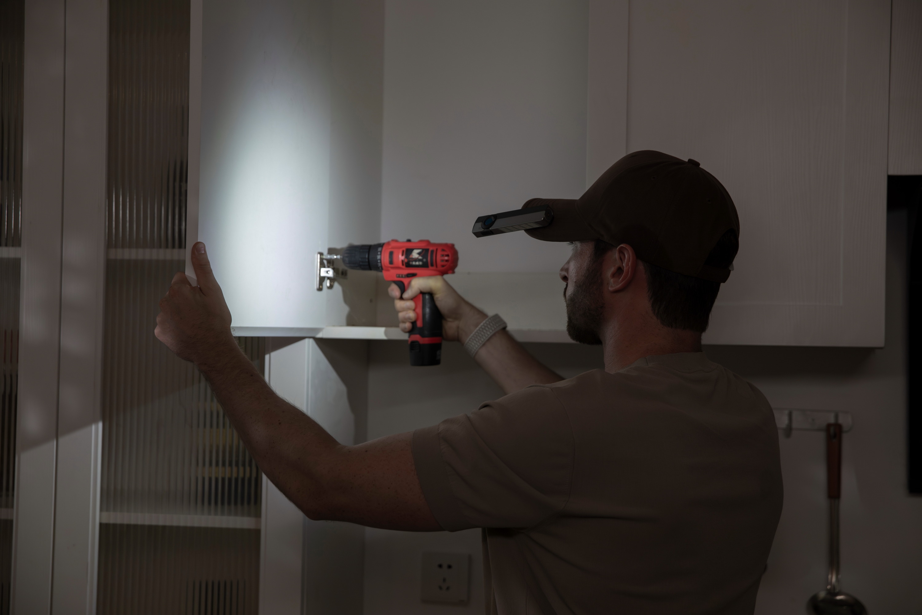 Handyman using a red cordless drill to install cabinet hinges, illuminated by a hands-free LED clip-on cap light for precision home repairs and DIY furniture assembly in low light.