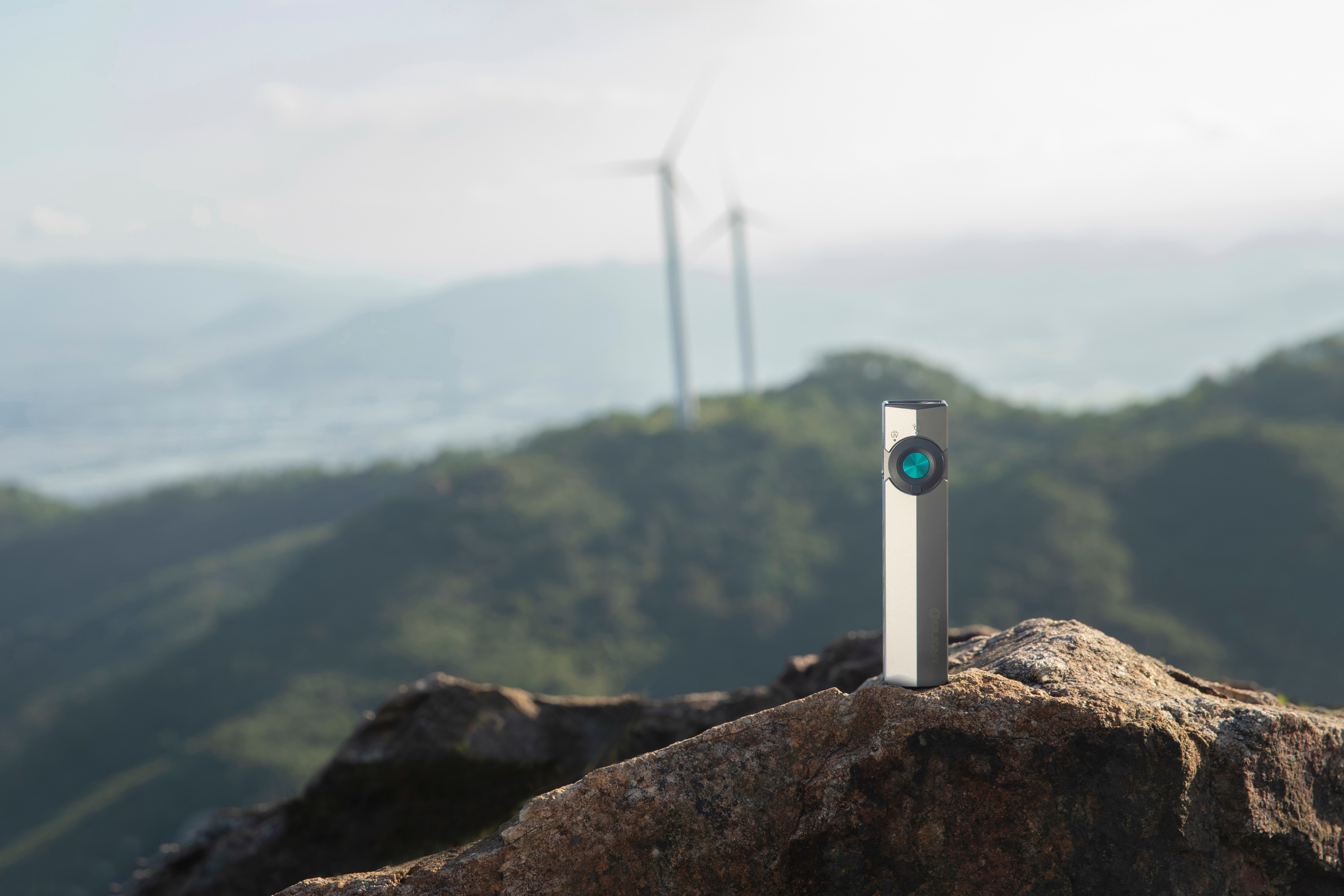 Compact silver EDC flashlight standing on a mountain rock with a scenic background of hills and wind turbines, ideal for hiking and outdoor adventures.