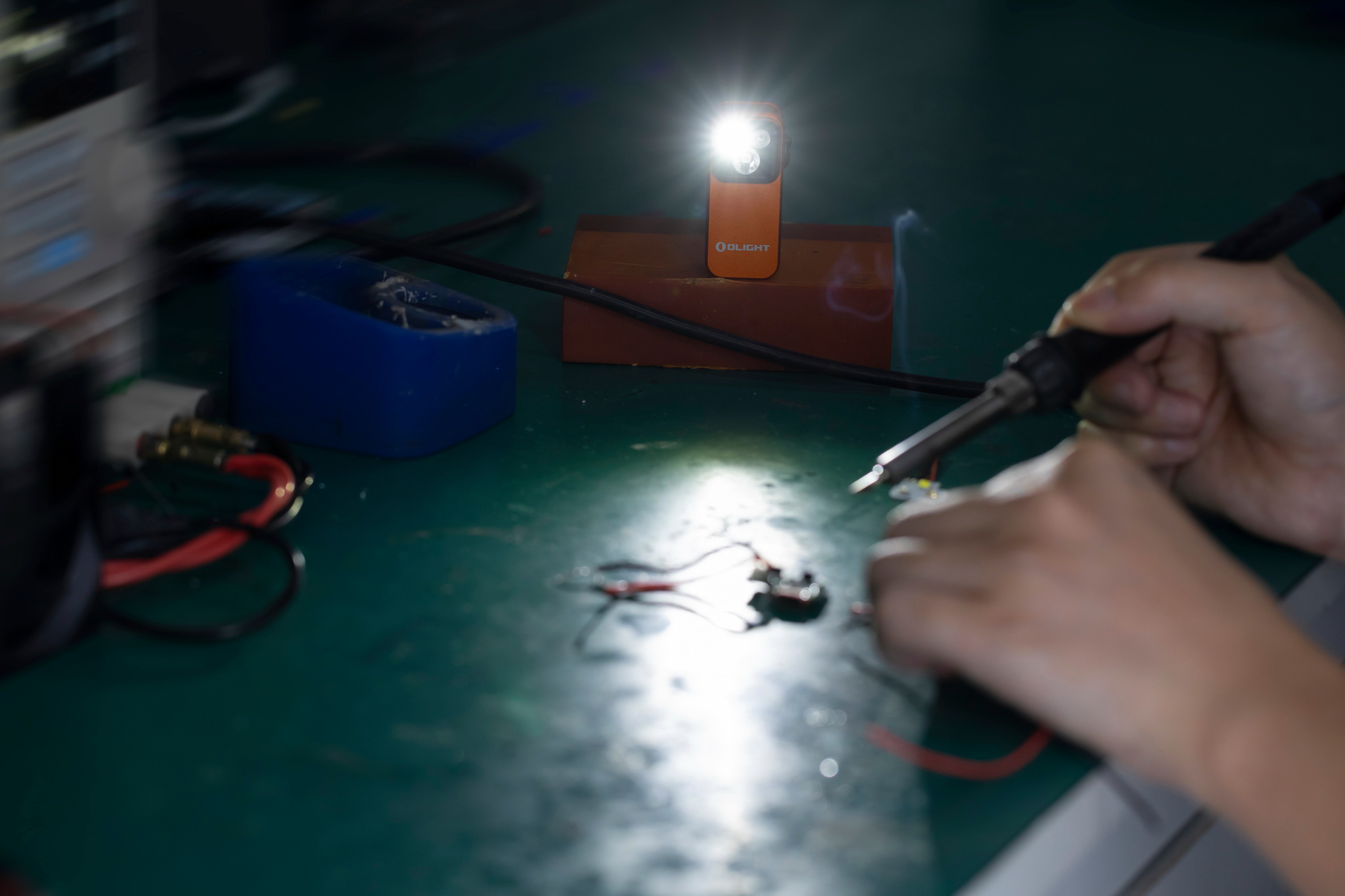 Orange Olight portable flashlight illuminating a person soldering electronic components on a workbench, providing bright focus for DIY repairs and precision work.