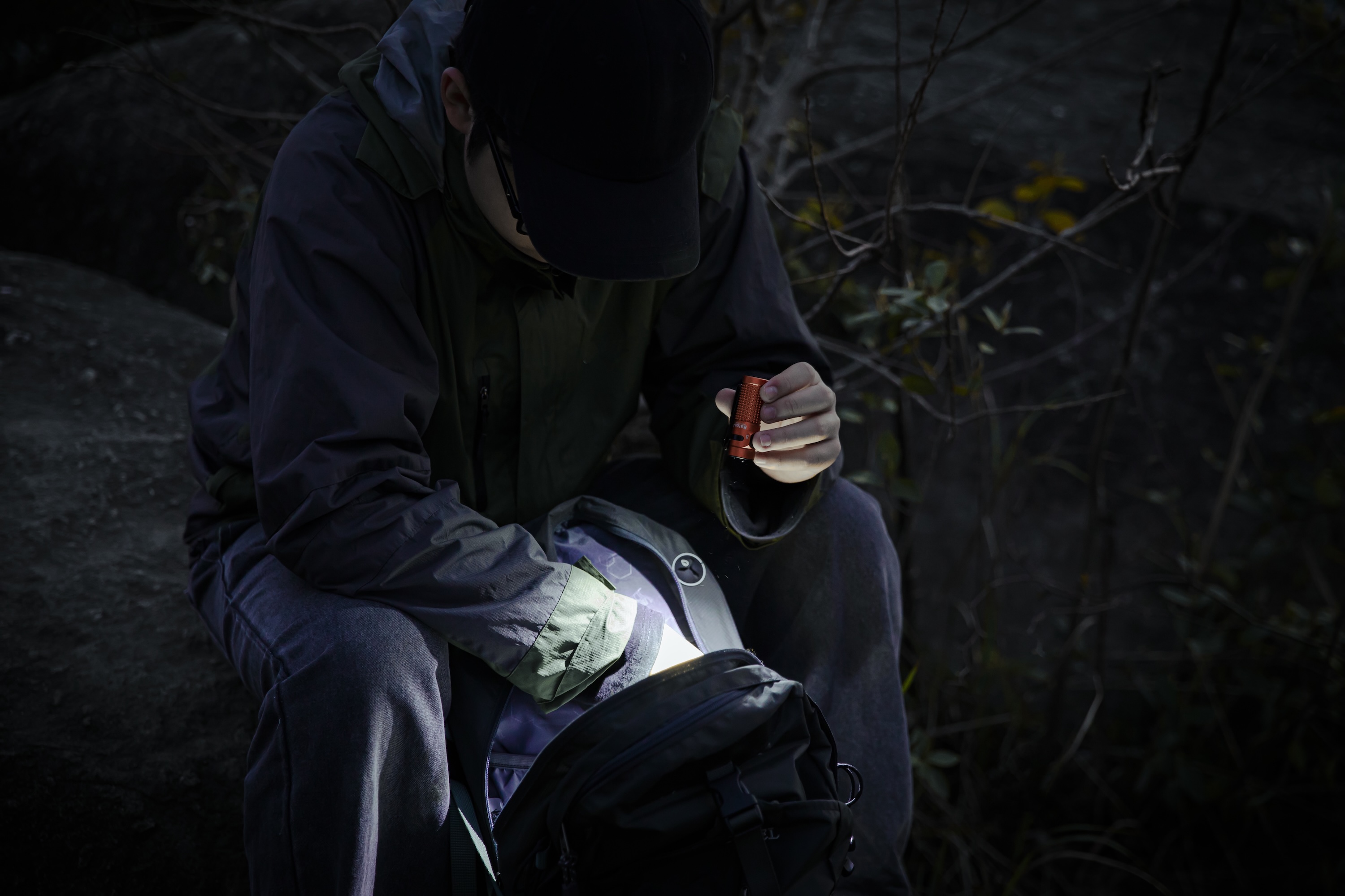 Man using a compact orange EDC flashlight to illuminate his backpack during a night outdoor activity, showcasing portable lighting solutions for hiking and camping.