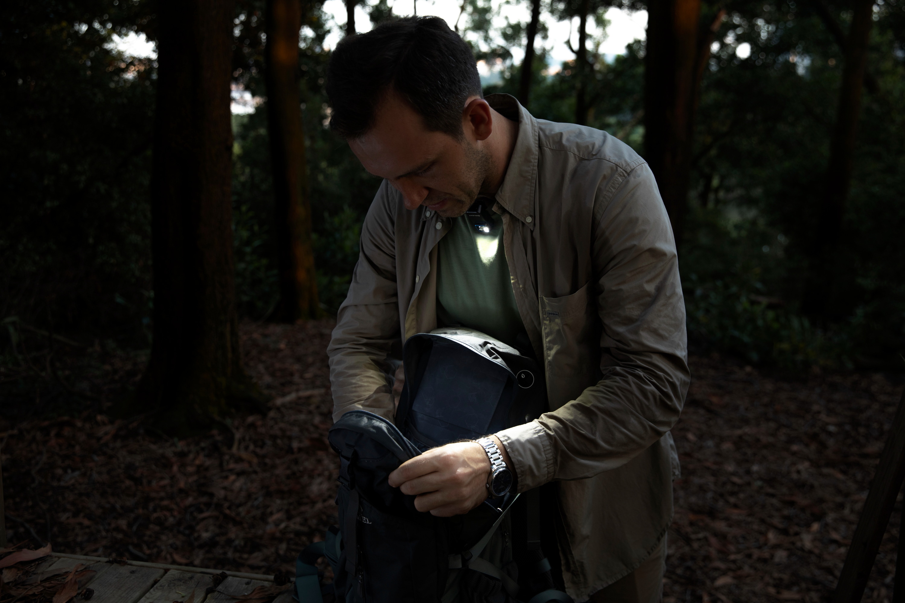 Man using a hands-free clip-on LED light to illuminate his backpack while hiking in a dark forest, showcasing portable outdoor lighting gear for camping and night adventures.