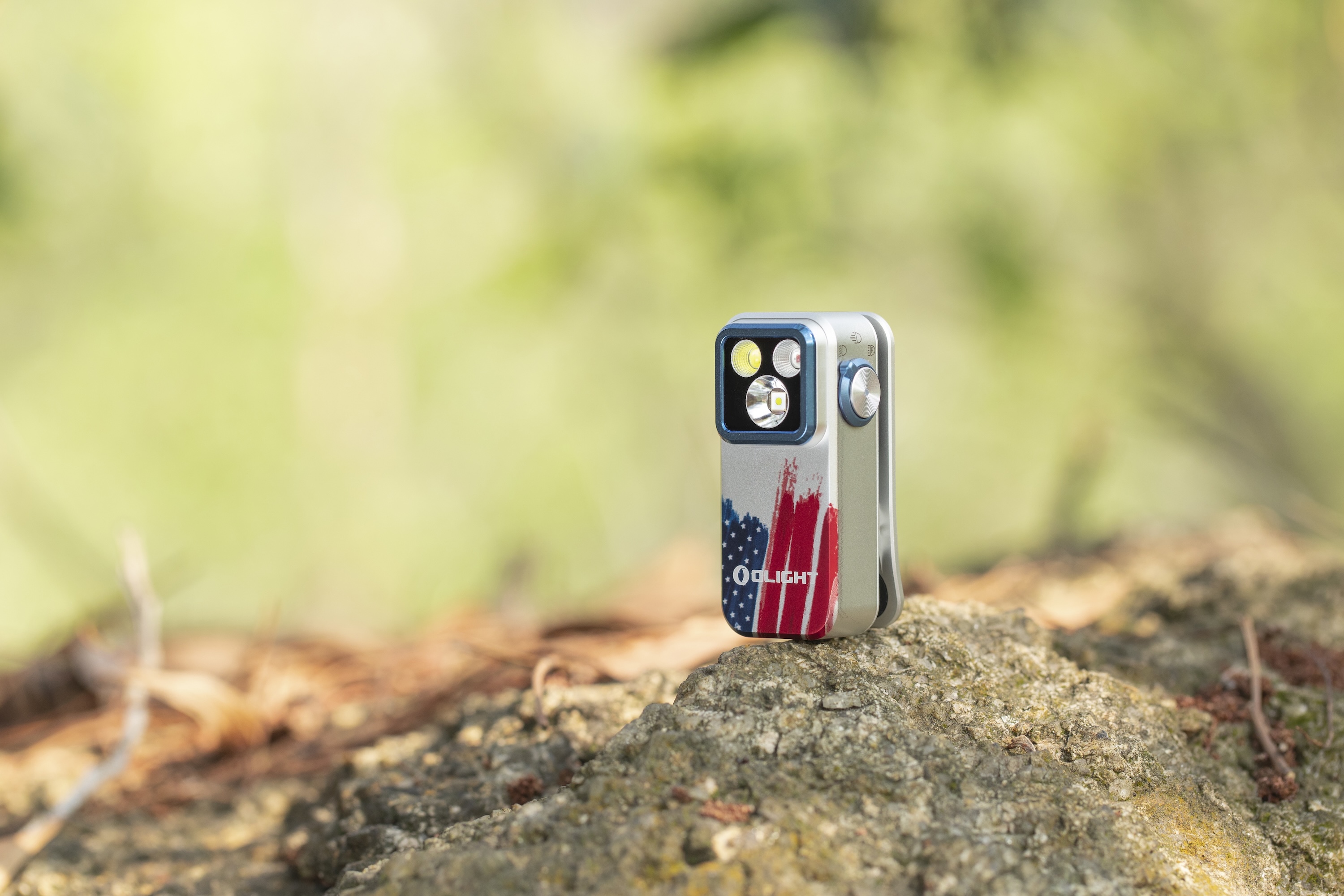 Lampe de poche Olight EDC compacte avec motif drapeau américain, posée sur un rocher en extérieur.