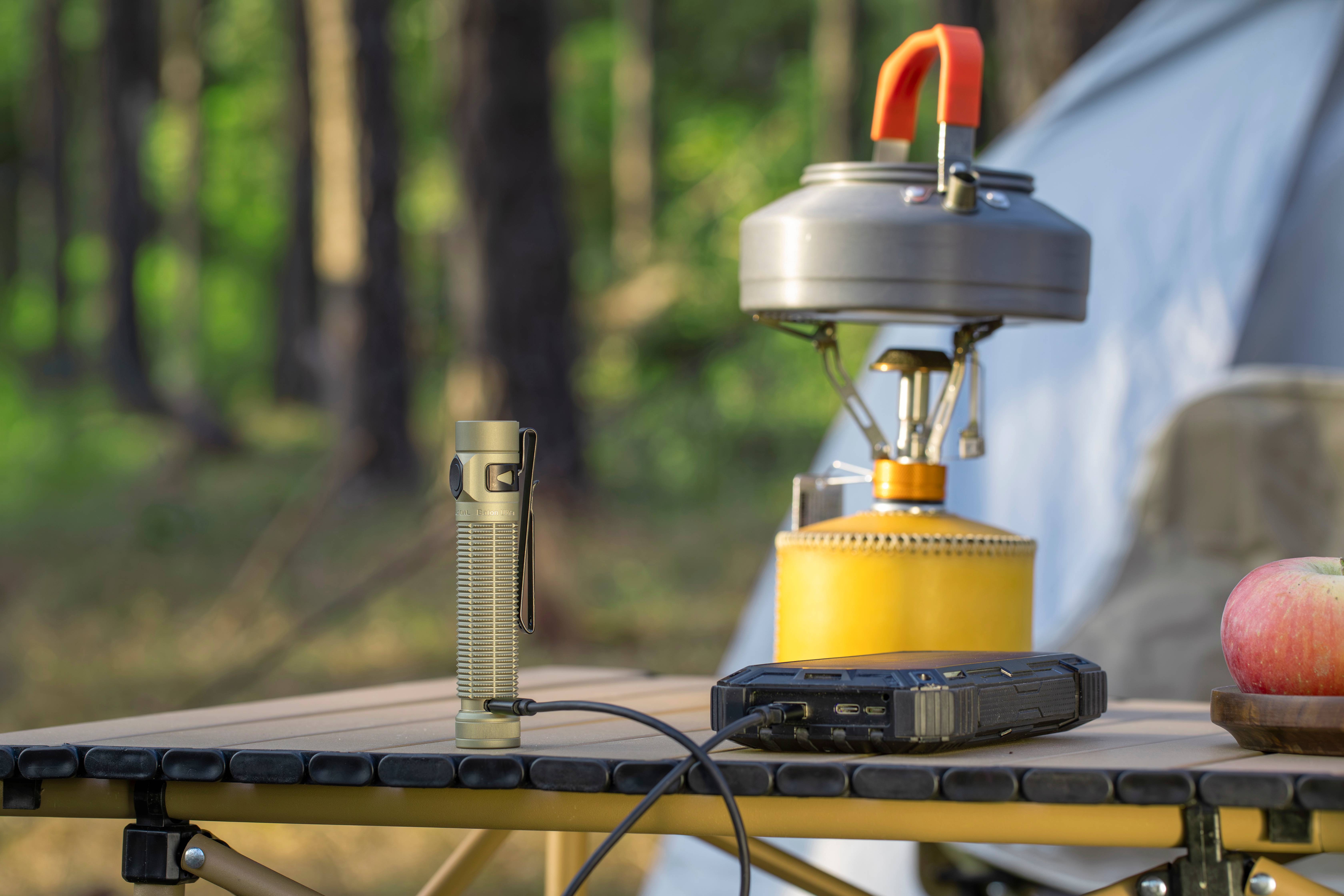 Tactical LED flashlight charging from a portable power bank on a camping table in the woods, with a camping stove, kettle, and tent in the background.