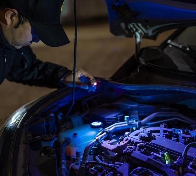 Man inspecting a car engine at night using a UV flashlight to check for leaks under the open hood.