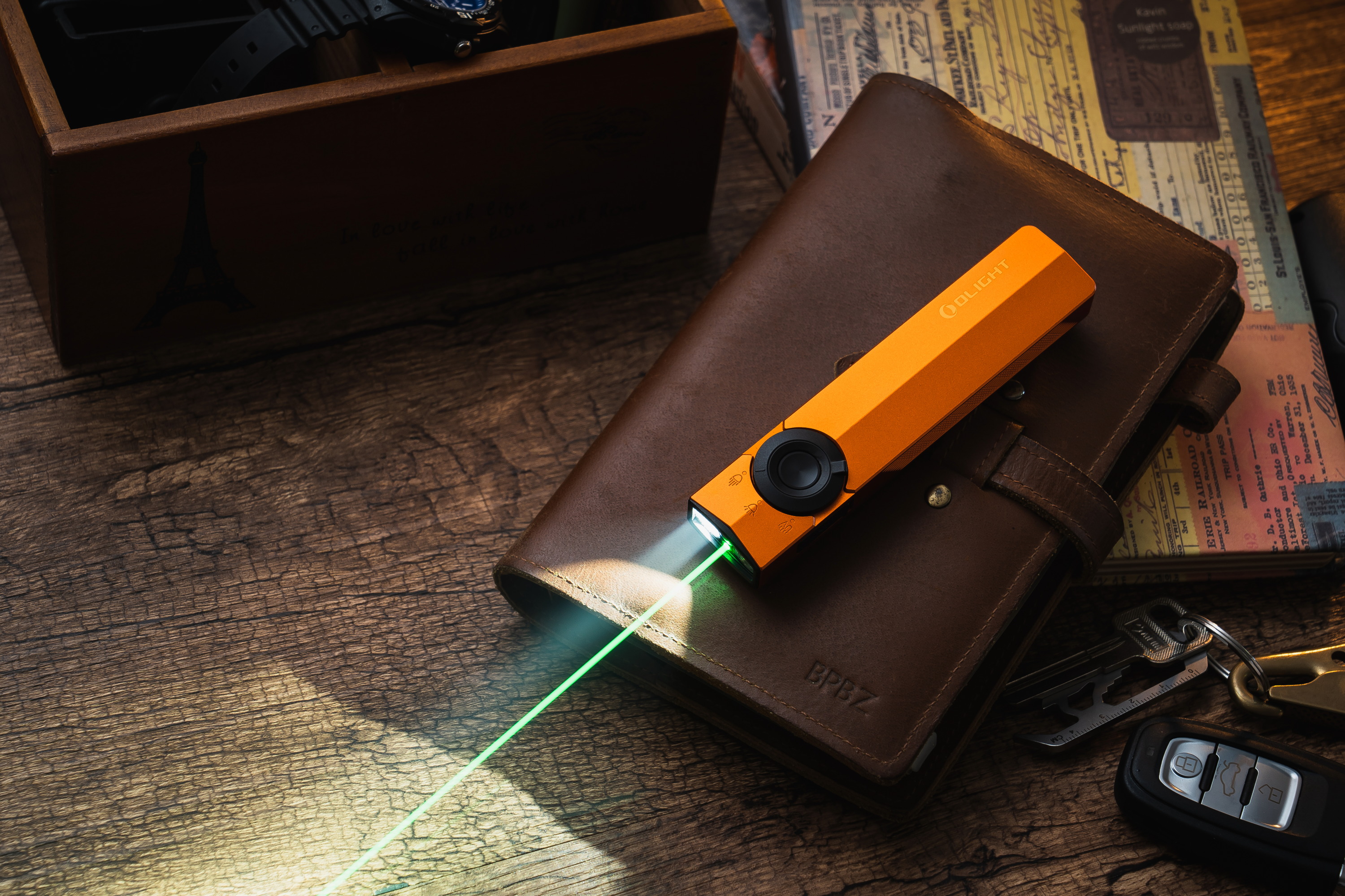 Orange Olight Arkfeld flat EDC flashlight with integrated green laser and white LED beam resting on a brown leather journal and wooden desk setup.