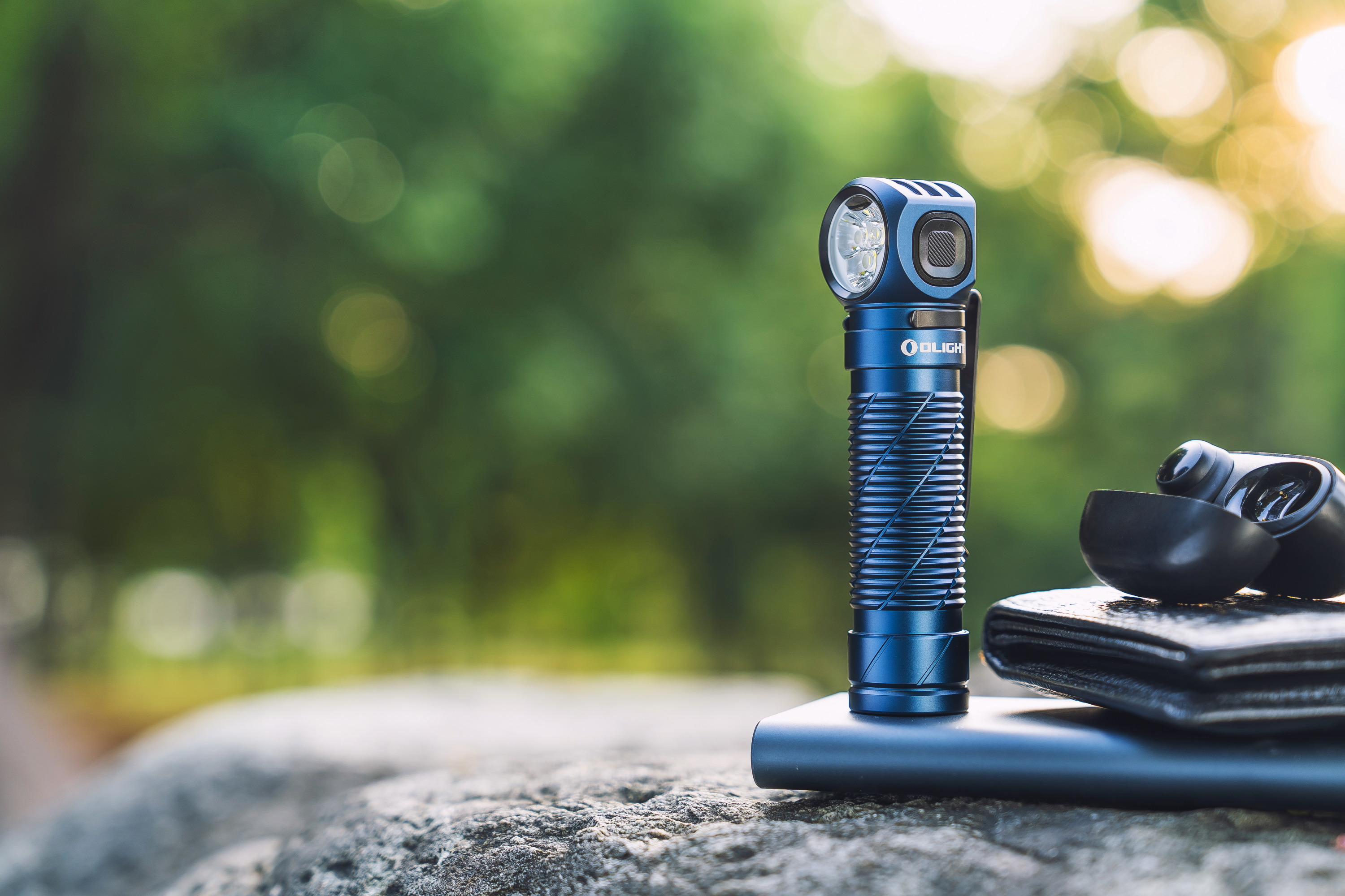 야외 바위 위에 놓인 파란색 올라이트 휴대용 손전등, 무선 이어버드, 지갑. 아웃도어 캠핑, 하이킹, EDC 필수품.