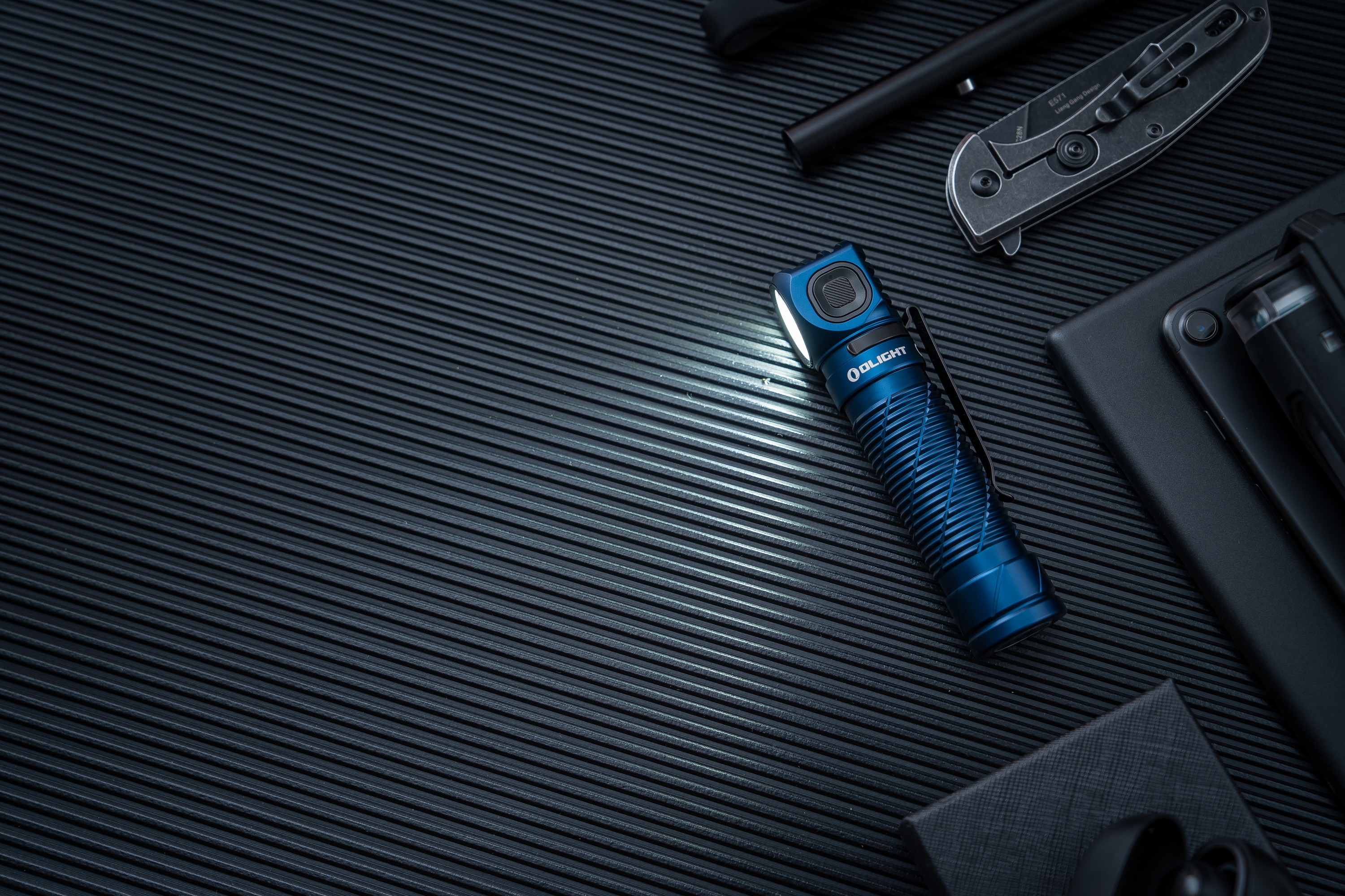 빛을 내는 파란색 Olight 손전등, EDC 나이프, 전술 장비가 검은색 배경에 놓여 있습니다.