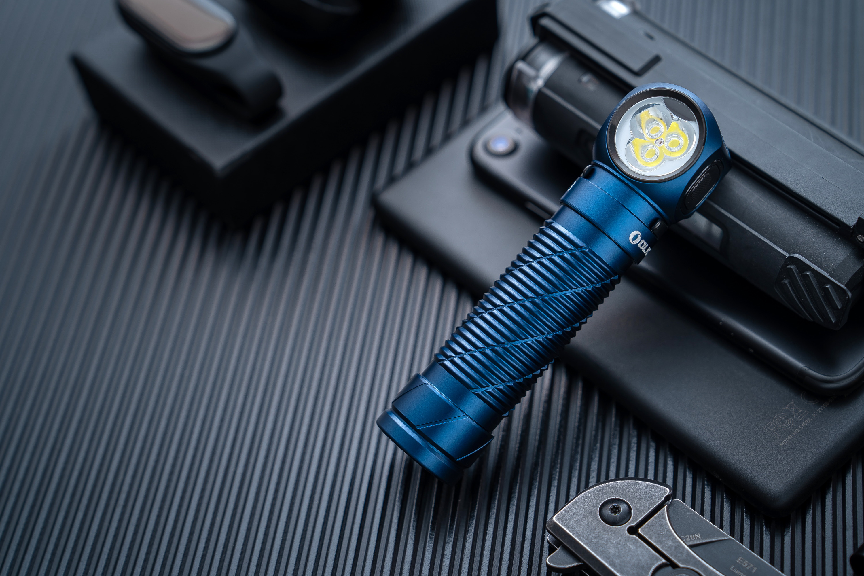 파란색 OLIGHT 손전등과 휴대폰, 나이프 등 일상 휴대 장비가 검은색 질감 배경 위에 놓여 있습니다.