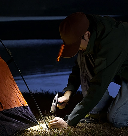 LED-Clip-Licht für Kappe beim Camping: Camper baut nachts ein Zelt auf und nutzt handfreie Beleuchtung an der Mütze, um Heringe sicher mit einem Hammer einzuschlagen.