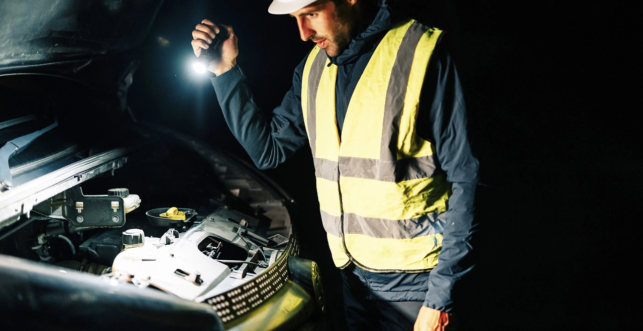 Roadside assistance worker in a yellow safety vest using a bright LED flashlight to inspect a broken-down car engine compartment at night.