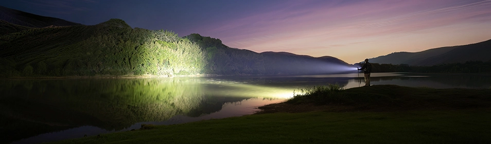 Lampe torche puissante LED &eacute;clairant un lac et une for&ecirc;t au cr&eacute;puscule. &Eacute;quipement de randonn&eacute;e nocturne avec un faisceau lumineux longue port&eacute;e haute intensit&eacute;.