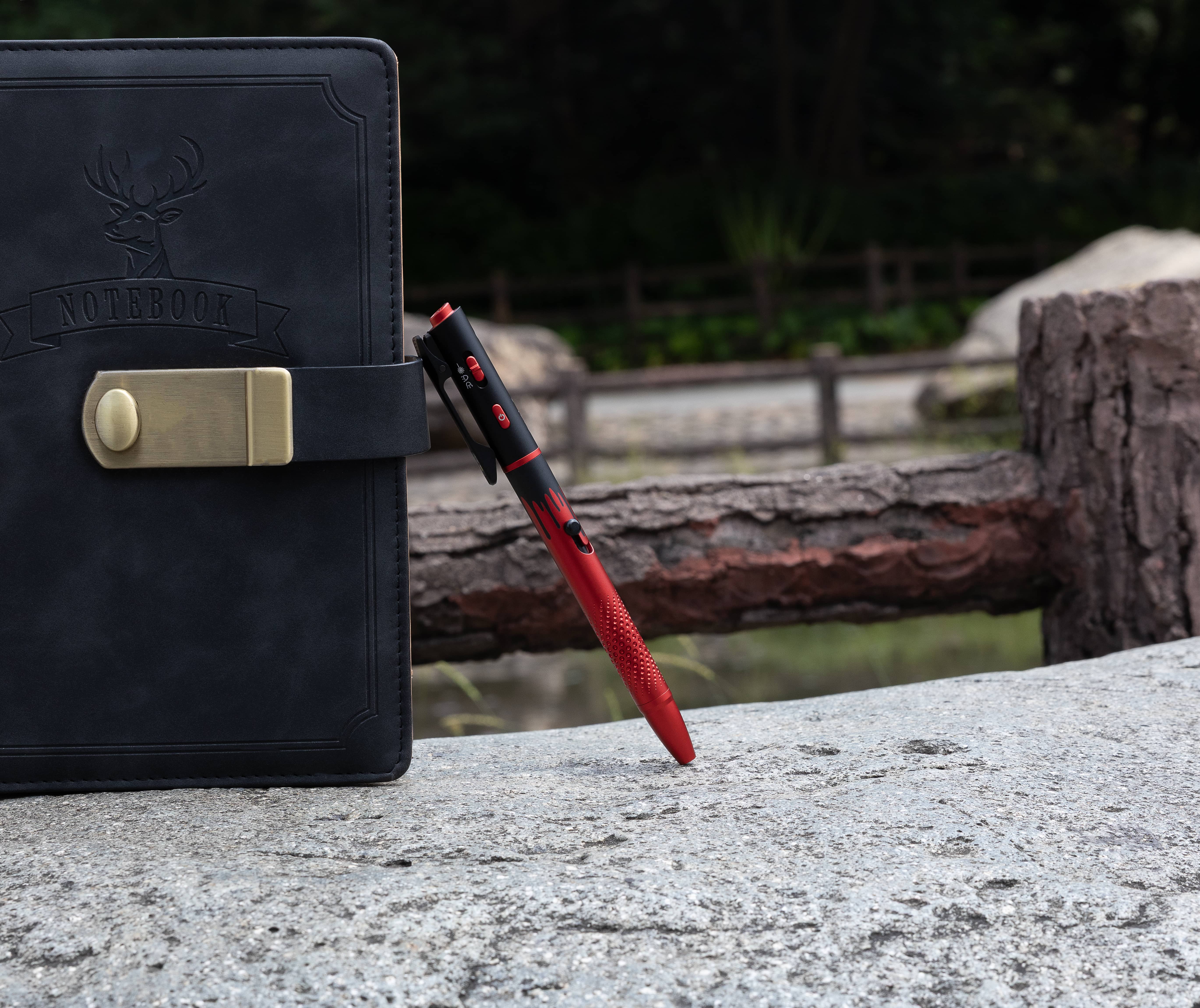 Black leather notebook with gold clasp and red tactical EDC pen on stone surface outdoors.