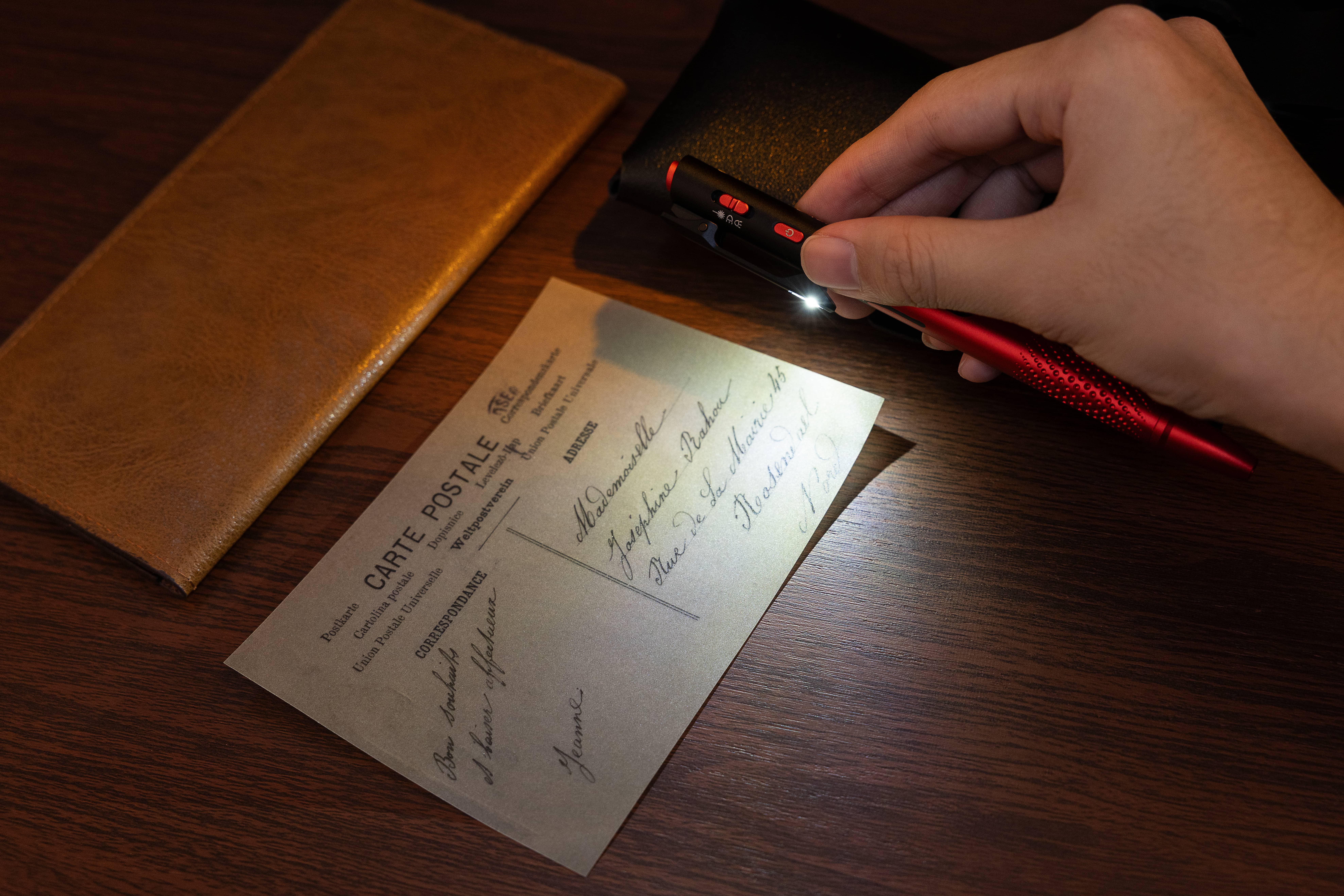 Hand holding a red EDC penlight with built-in LED flashlight illuminating a vintage postcard on a wooden desk.
