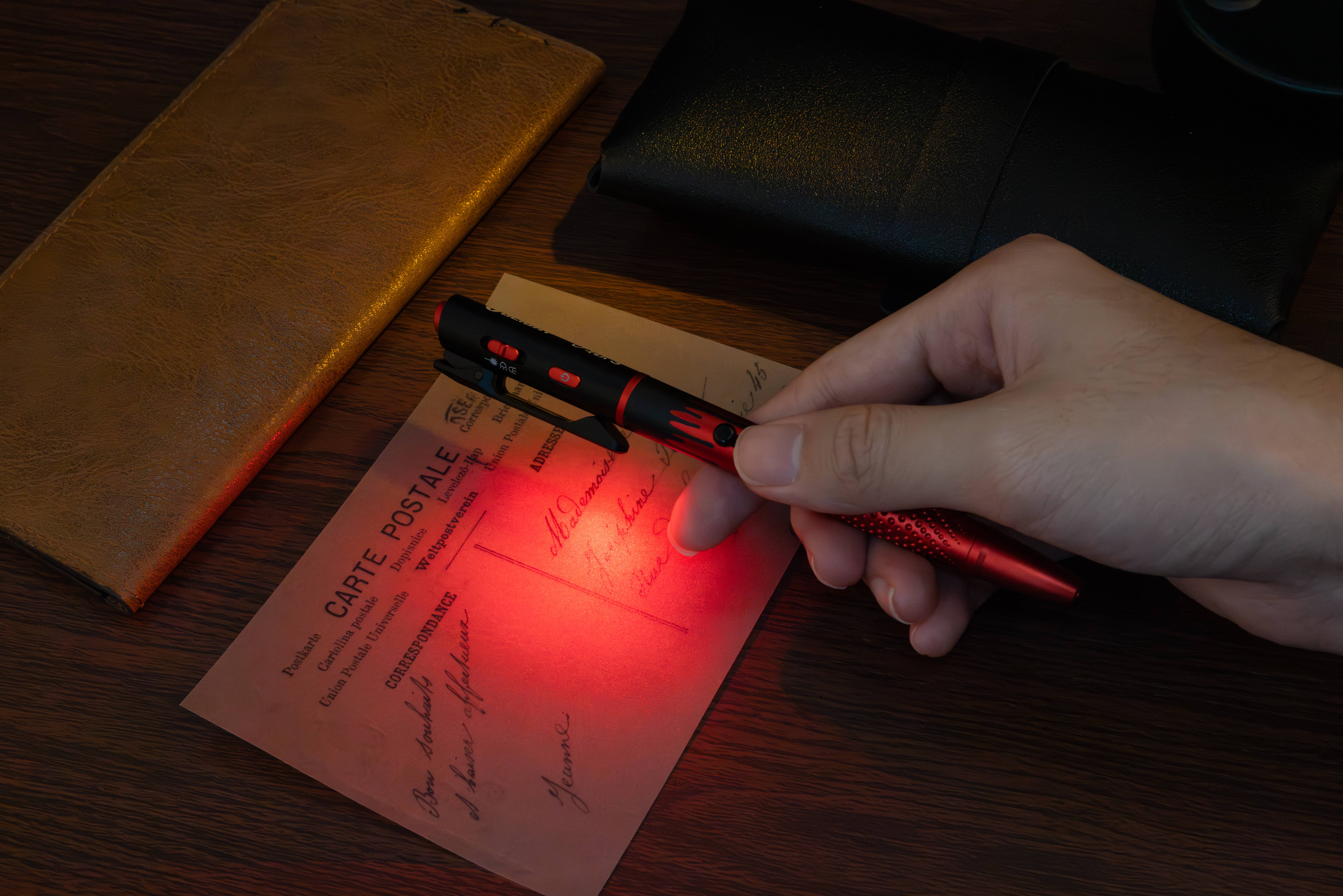 Tactical pen flashlight illuminating a vintage postcard with red light on a wooden desk.