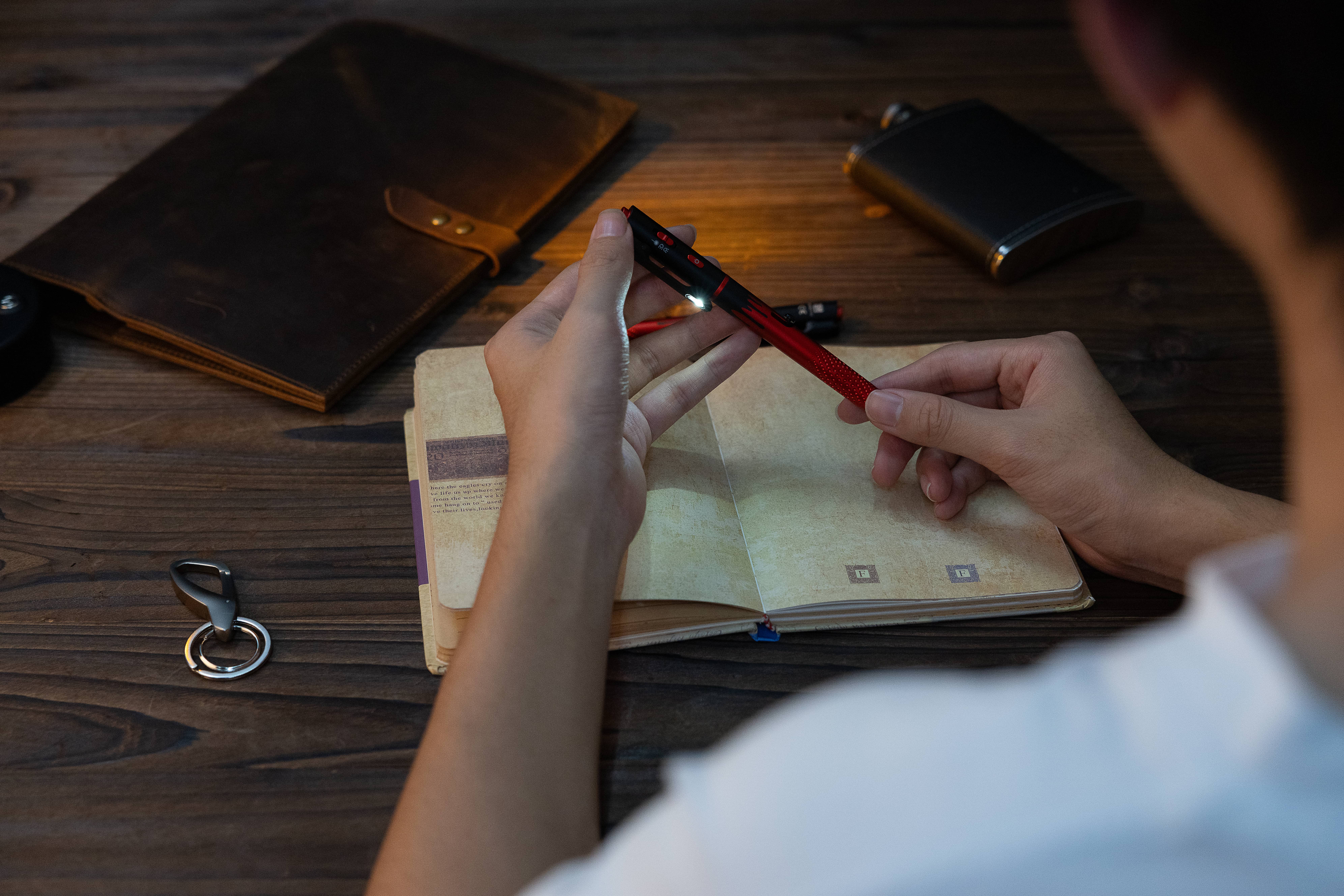 Hand holding a red EDC pen flashlight illuminating a vintage journal on a rustic wooden desk with leather notebook
