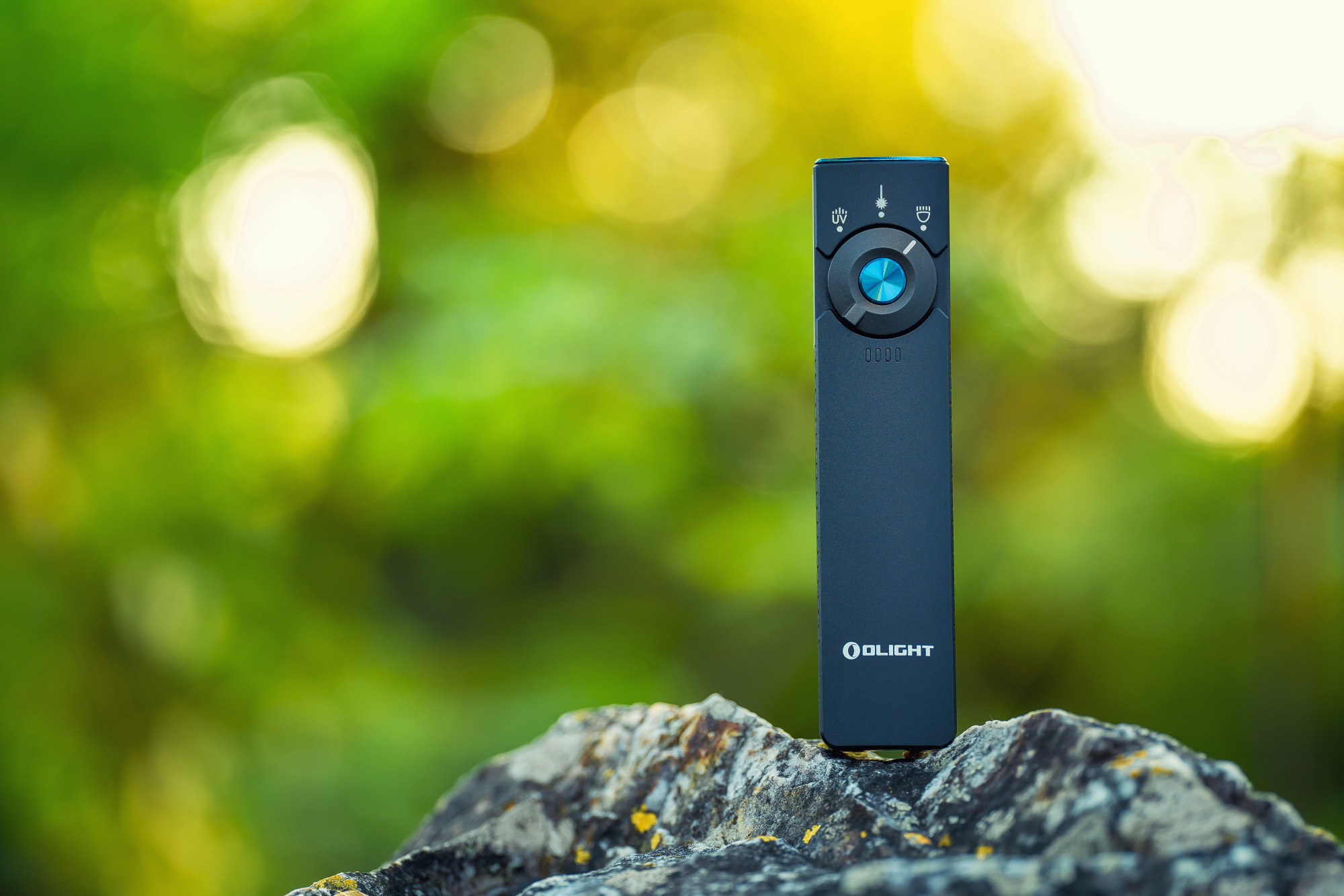 OLIGHTのブラックな懐中電灯、屋外で岩の上に設置され、鮮やかな緑と黄色の背景にぼやけた木々がある。