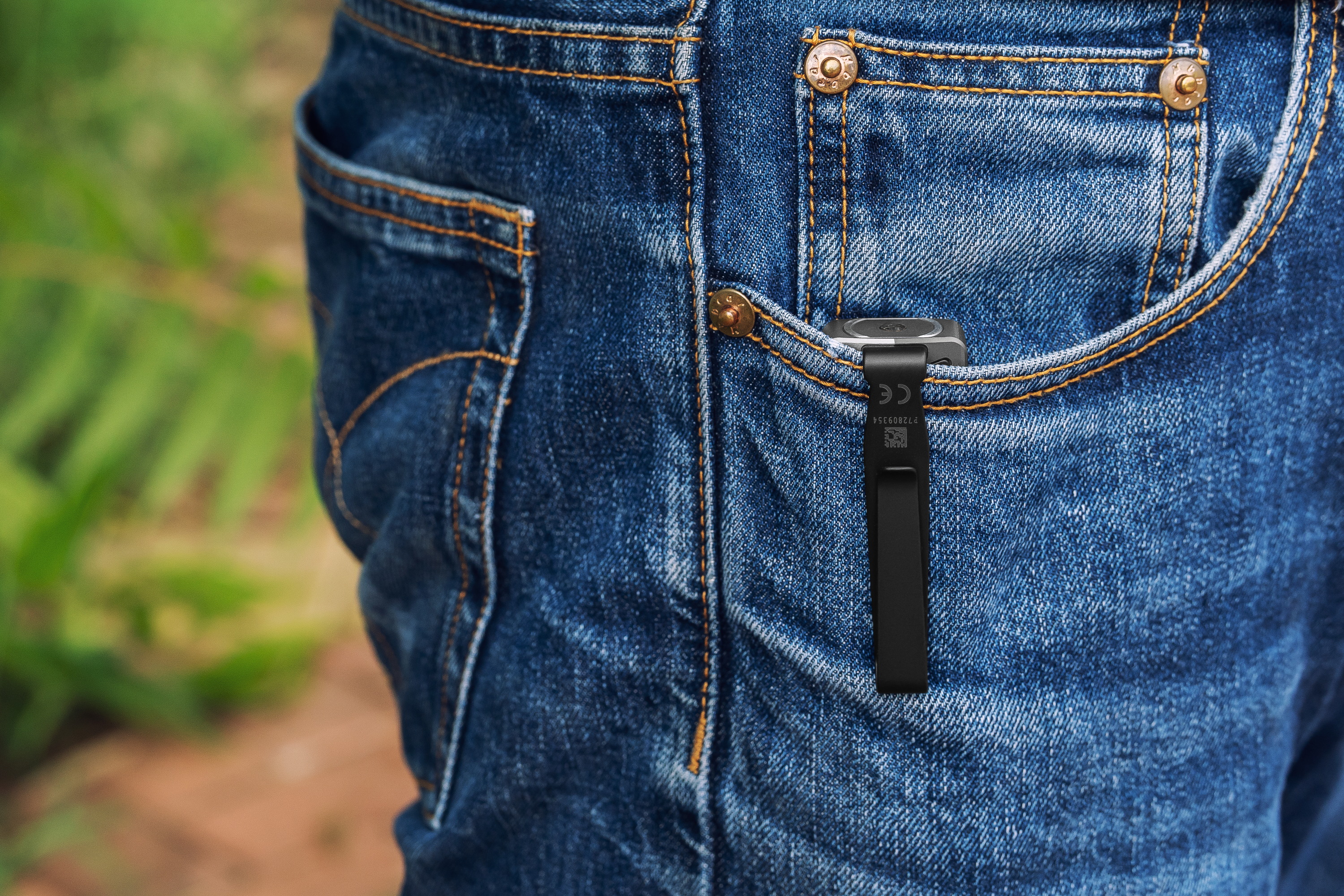 Black clip-on portable device, possibly an audio recorder or tracker, secured in a blue denim jeans pocket.