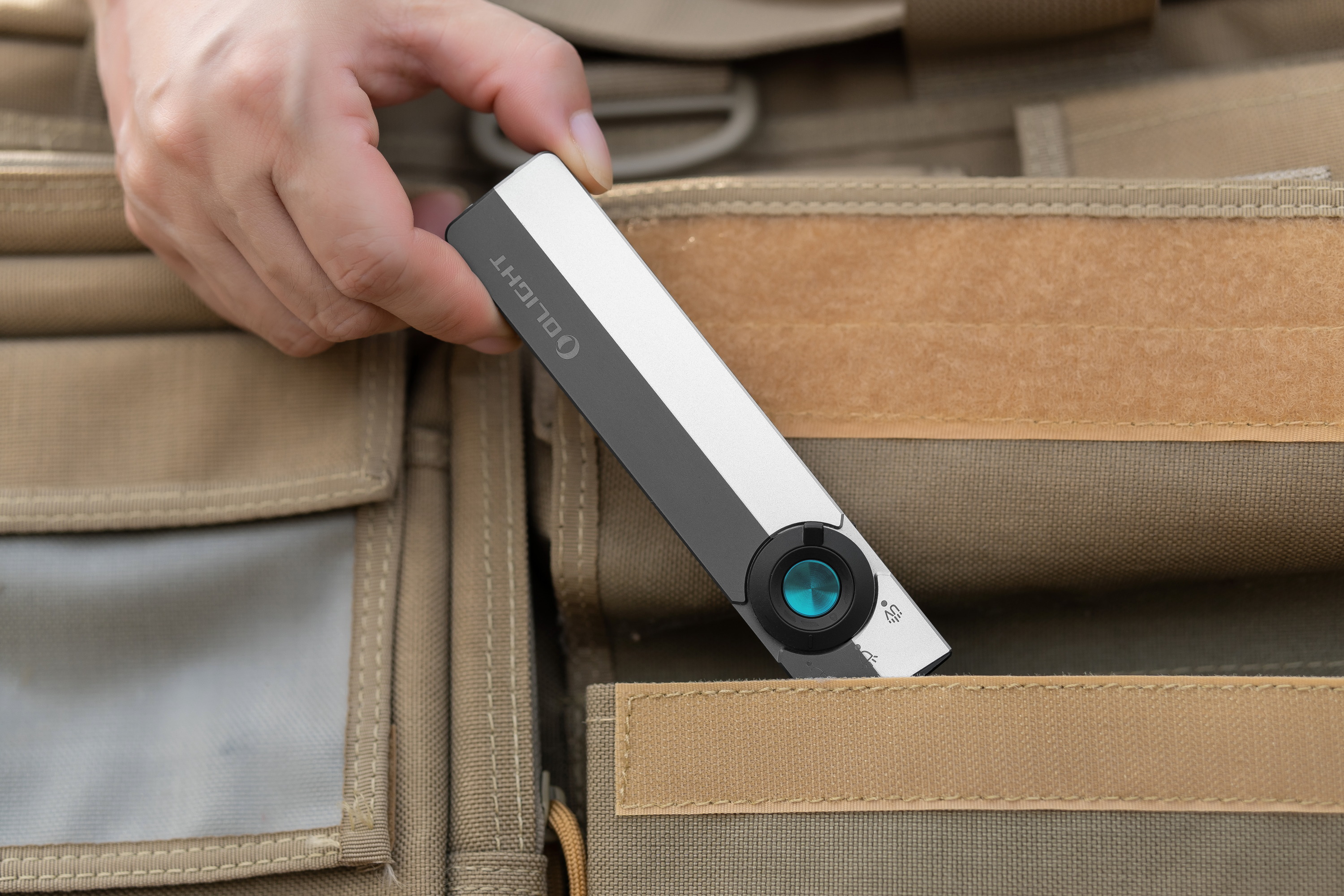 Hand placing a slim Olight Arkfeld flat EDC flashlight into a tan tactical bag pocket, highlighting its compact design and portability for outdoor and daily use.