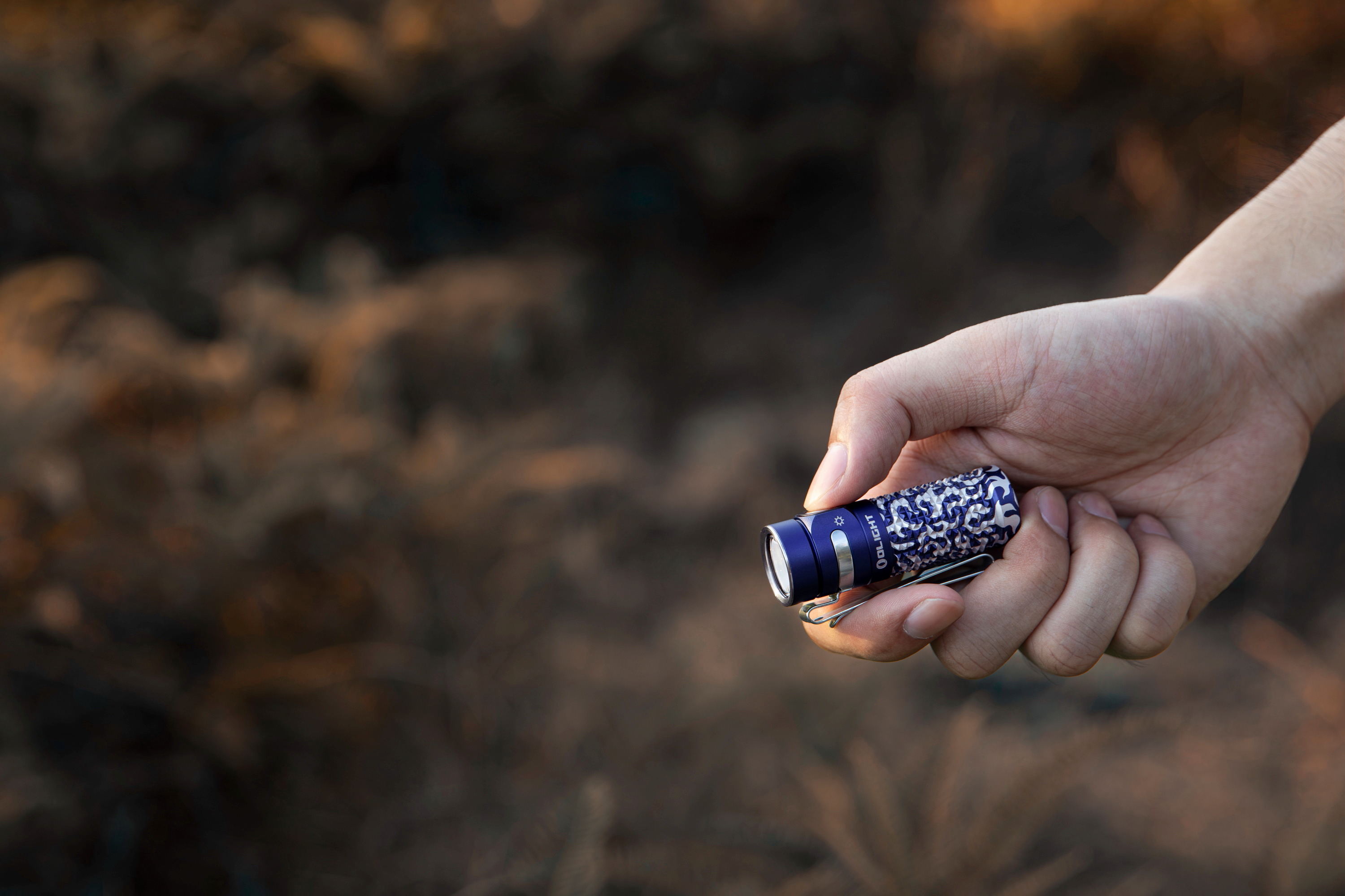 Main d'un homme tenant une mini lampe de poche EDC Olight compacte avec motif camouflage bleu et clip de poche en acier, id&eacute;ale pour le camping et la randonn&eacute;e en plein air.