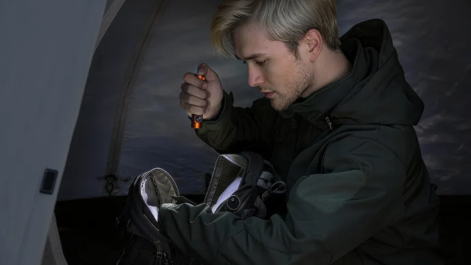 Homme utilisant une lampe torche LED compacte pour chercher dans son sac &agrave; dos sous une tente de camping la nuit.