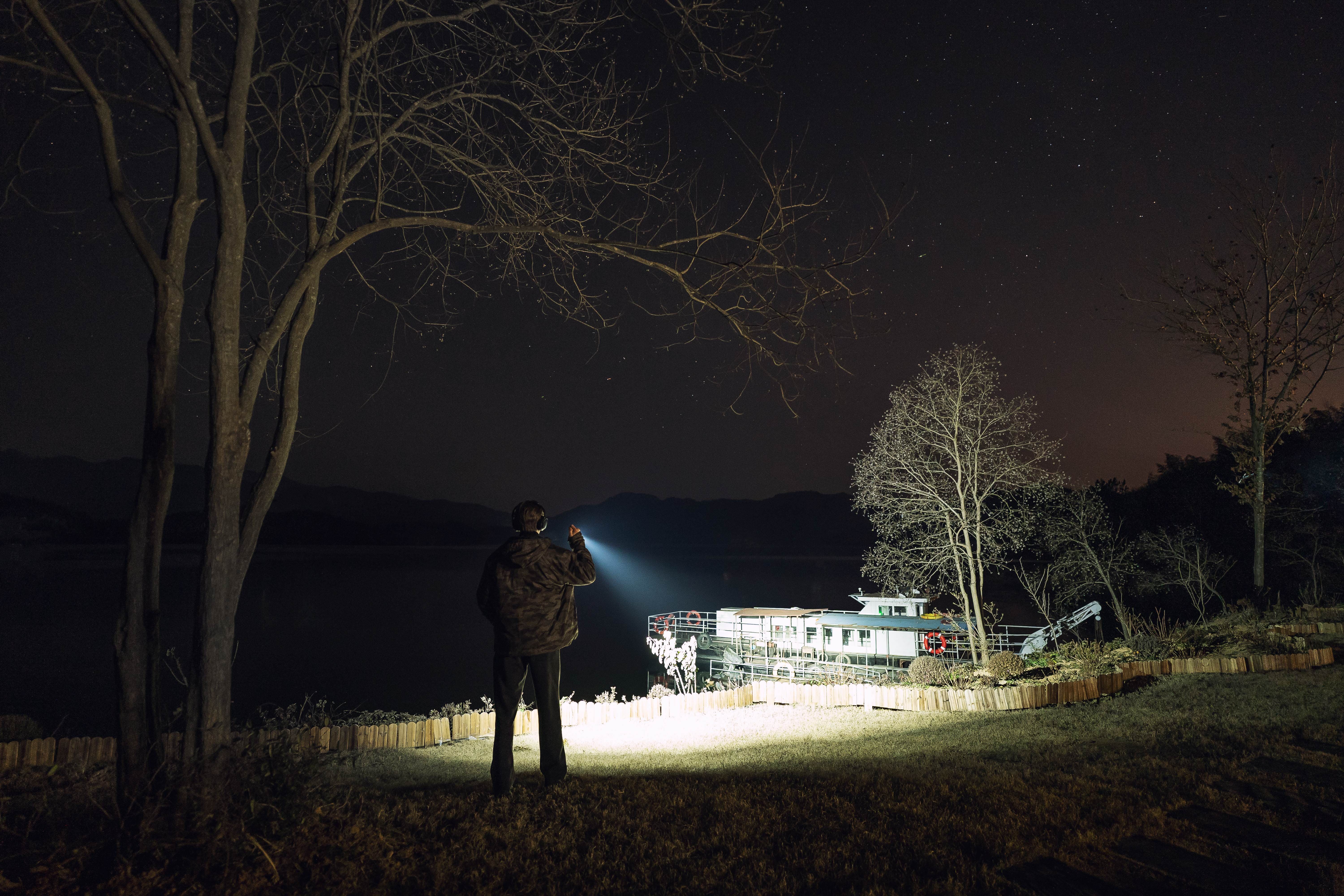 A man standing on a grassy hill at night, shining a powerful LED tactical flashlight towards a boat on a lake under a starry sky.
