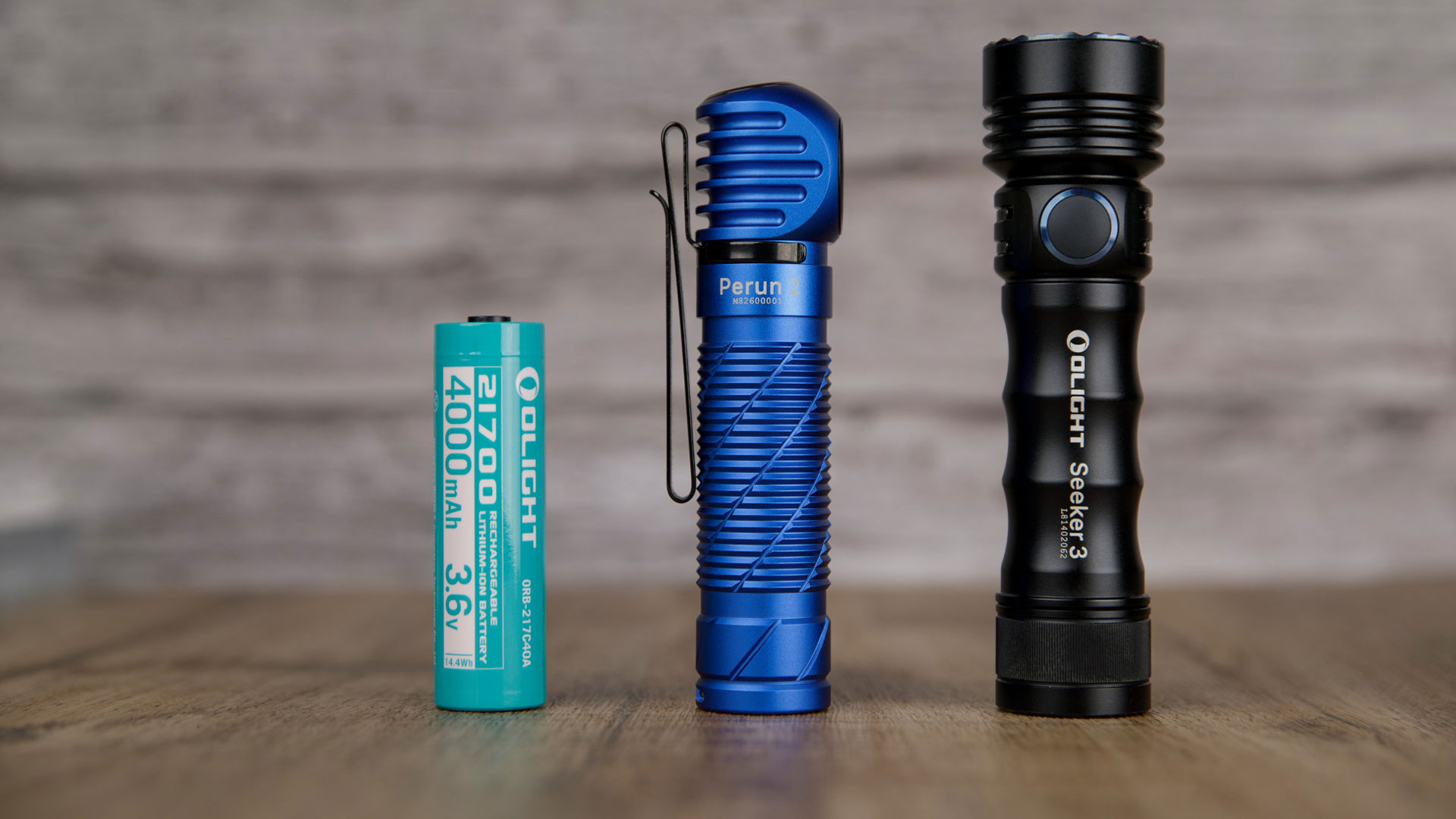 Comparaison de taille entre la batterie 21700 Olight, la lampe frontale coud&eacute;e Perun 2 bleue et la lampe torche Seeker 3 noire sur table en bois