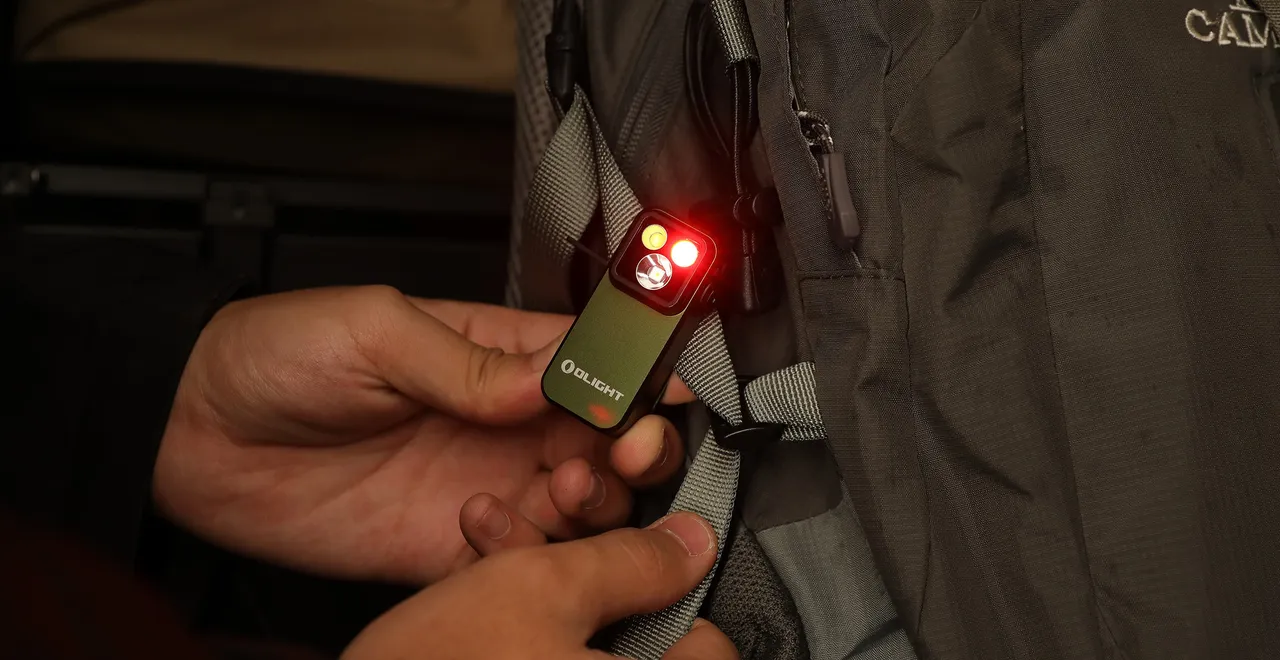 Olight clip light in olive green with active red and white beams being secured to a hiking backpack strap.