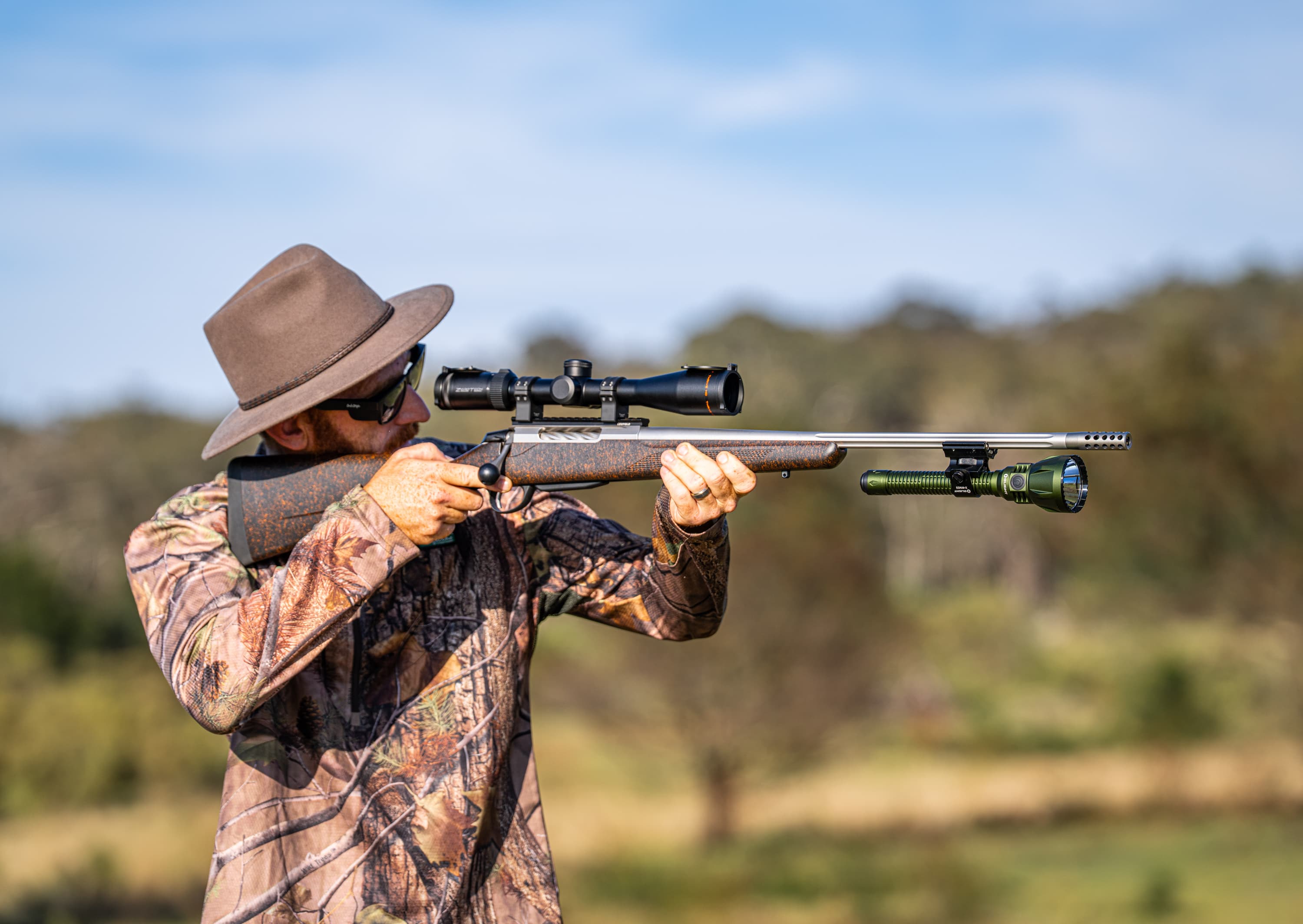 Hunter with rifle, scope, and attached flashlight aims in a natural outdoor setting