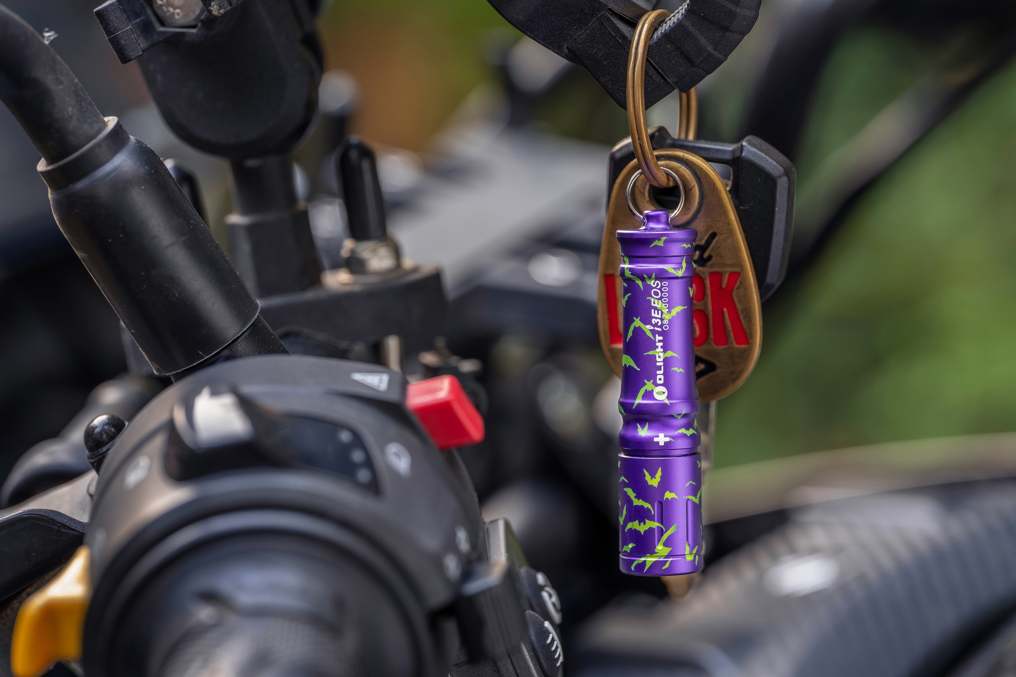 Mini lampe torche porte-cl&eacute;s Olight i3E EOS violette avec motifs chauves-souris vertes, suspendue au guidon d'une moto avec des cl&eacute;s.