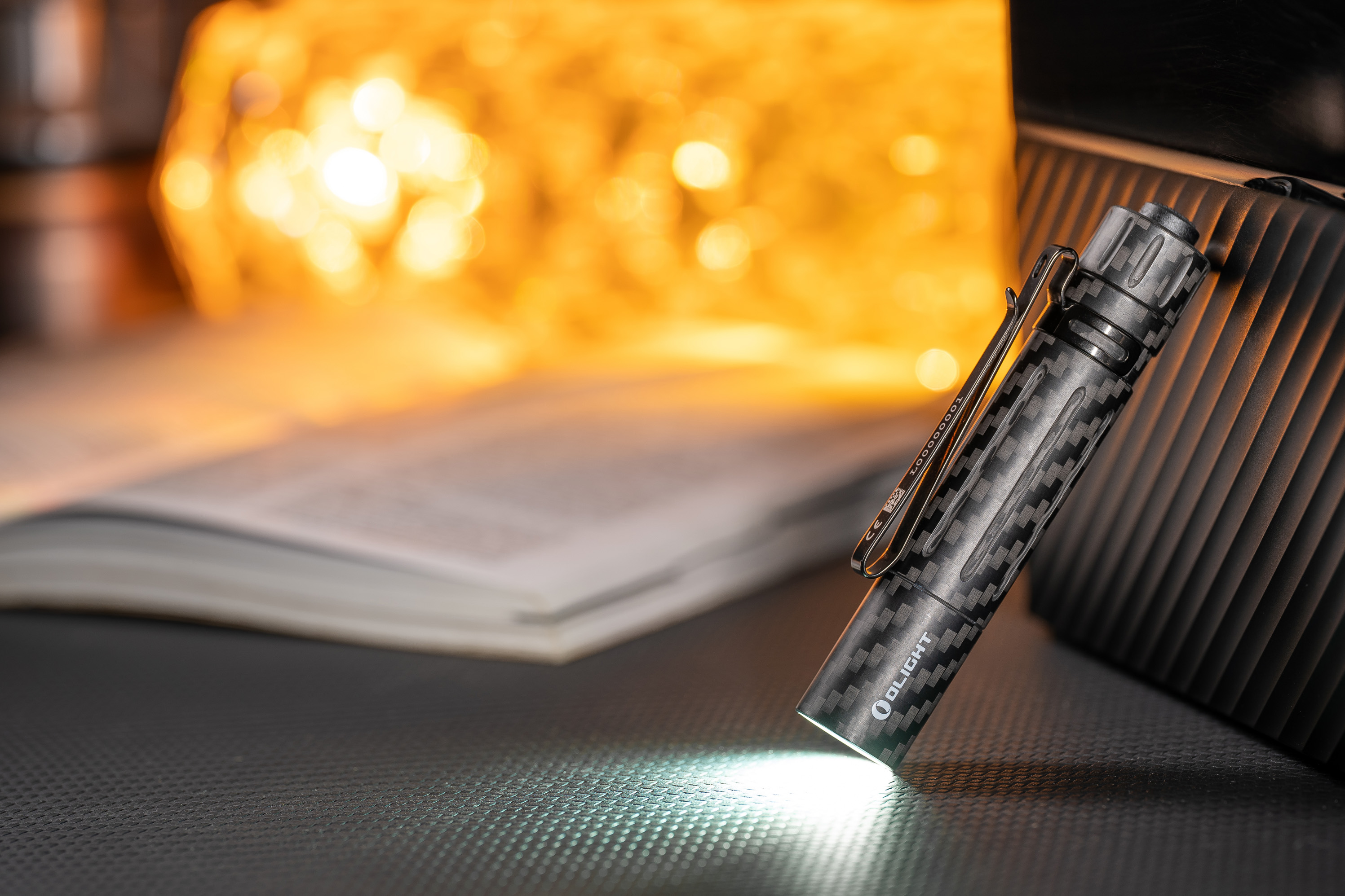 Lampe torche EDC Olight en fibre de carbone allum&eacute;e sur un bureau avec livre et &eacute;clairage chaleureux en arri&egrave;re-plan