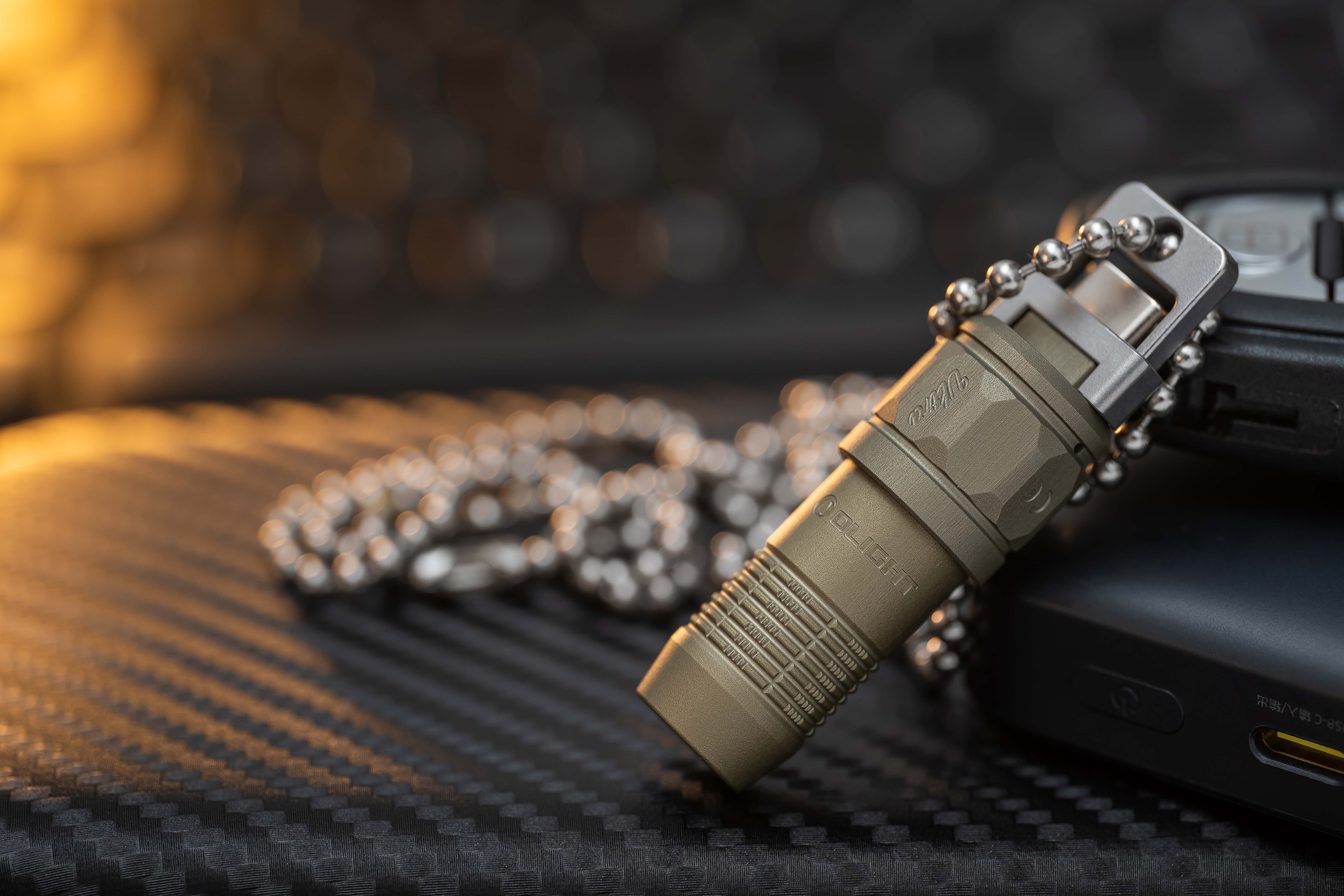 Linterna Olight táctica de color caqui con cadena, conector USB-C y logotipo "Ulite", sobre una superficie texturizada. Ideal para equipo EDC.