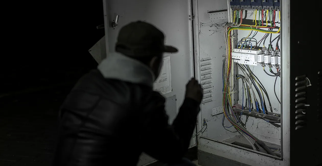 T&eacute;cnico electricista inspeccionando y reparando un cuadro el&eacute;ctrico y cableado industrial de noche con una linterna.