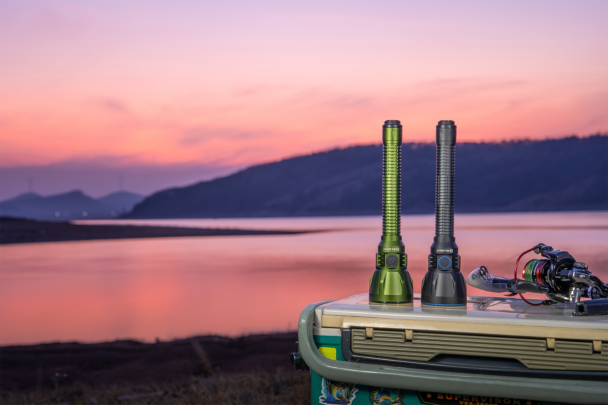 Two flashlights, green and black, on a cooler beside a lake with a fishing reel at sunset