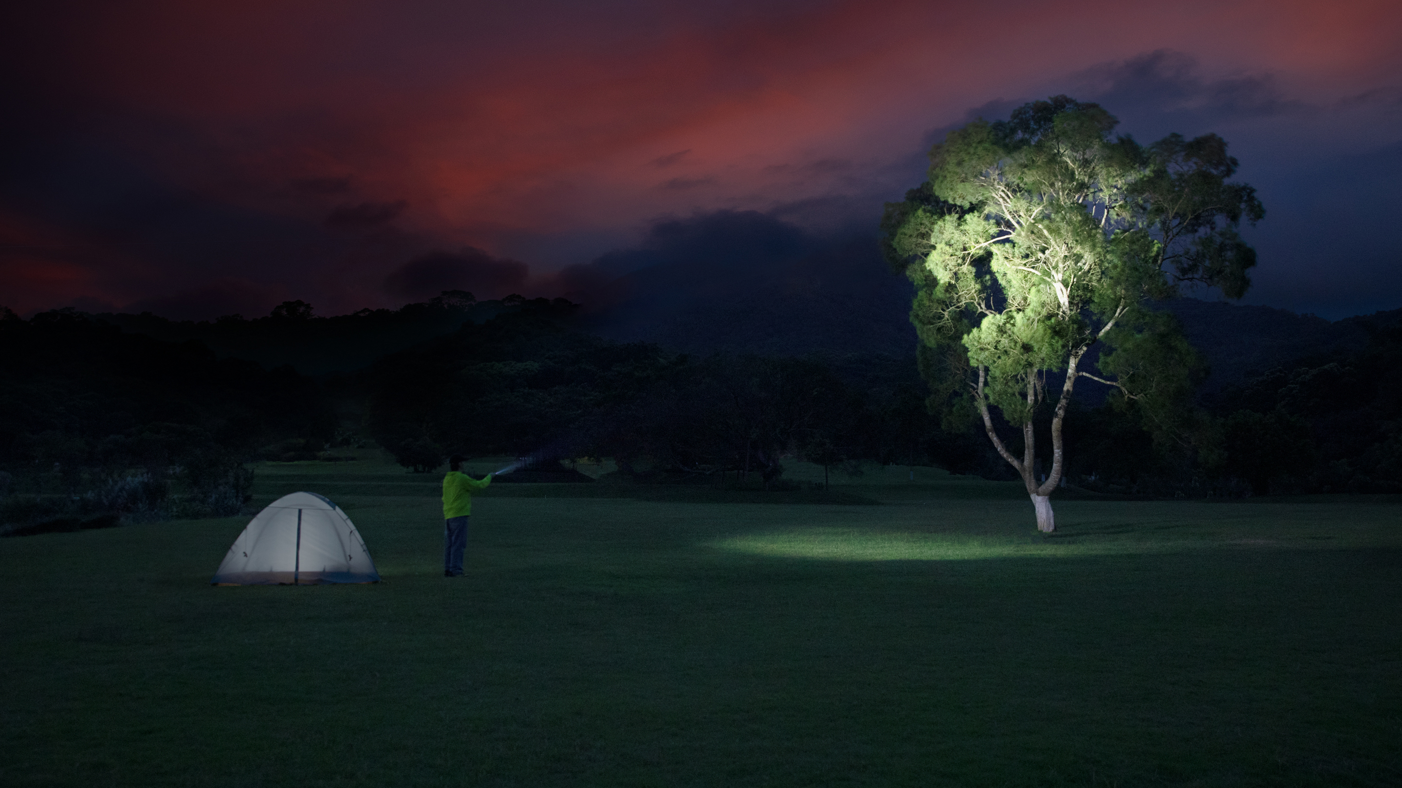 Man camping in a green field at night, shining a bright flashlight on a large tree next to his illuminated tent under a dramatic red and dark sunset sky.