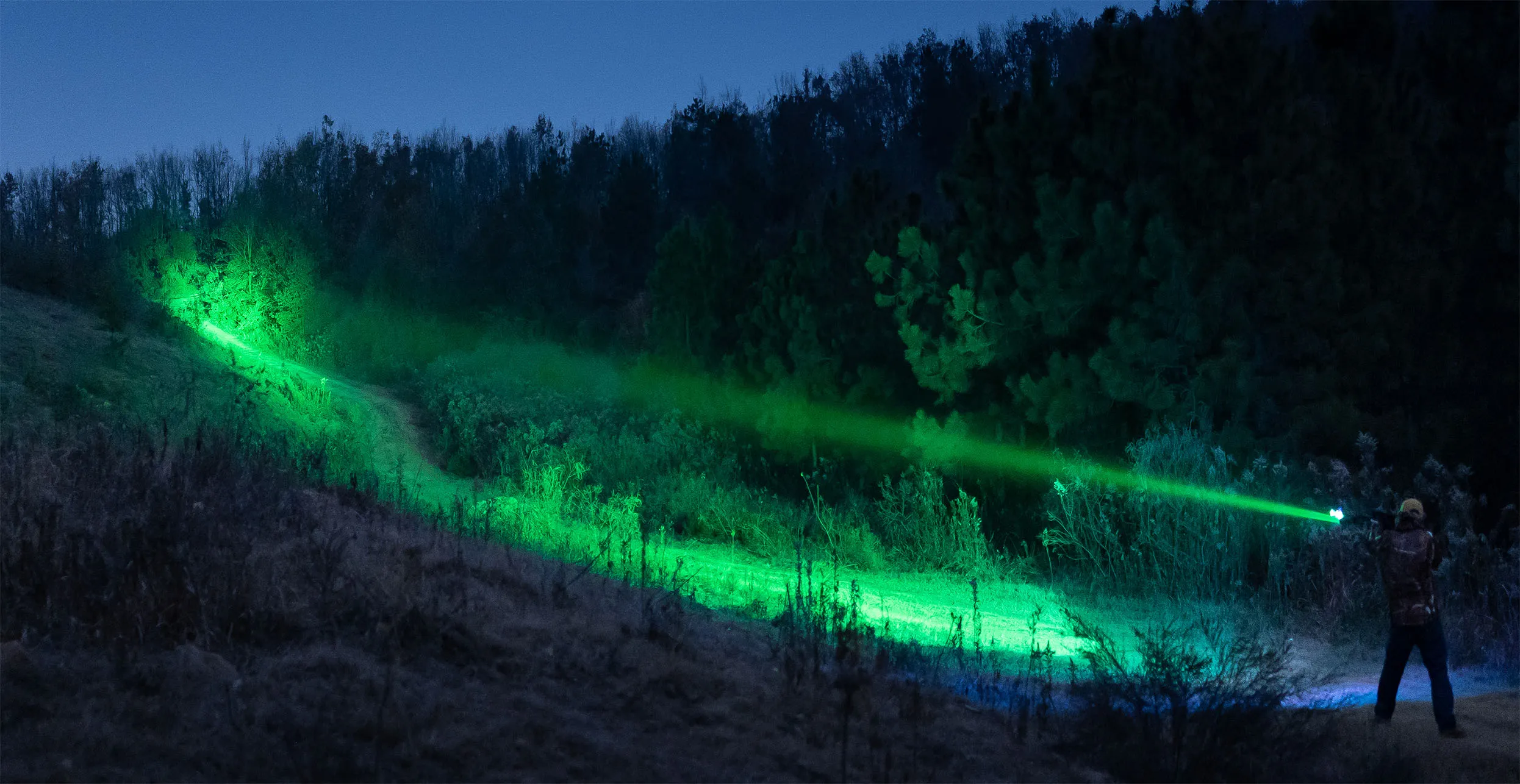 Hunter using a high-power green tactical flashlight beam to illuminate a forest trail at night