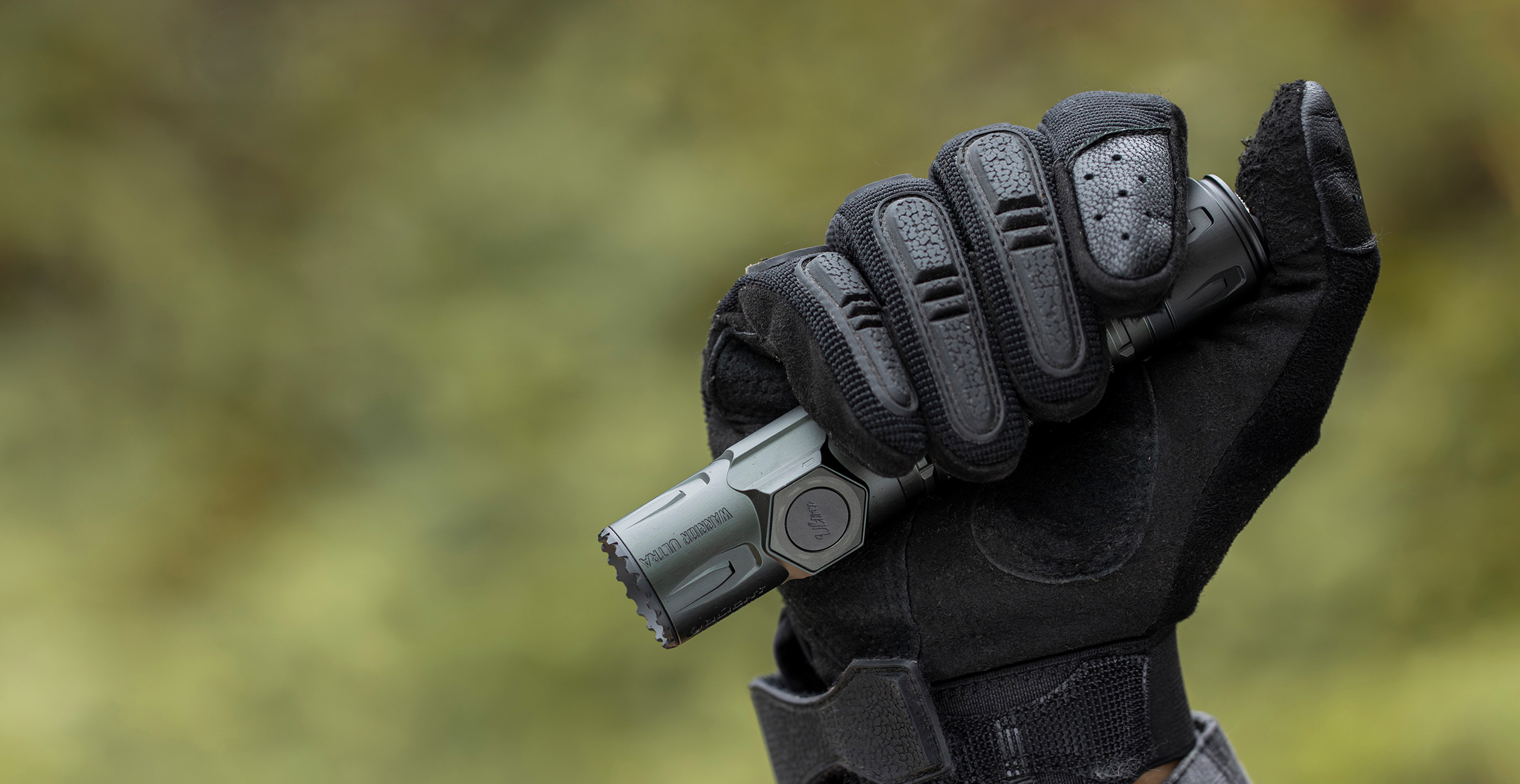Hand in taktischen Handschuhen hält eine robuste Outdoor-Taschenlampe (Warrior Ultra) vor verschwommenem Grün.