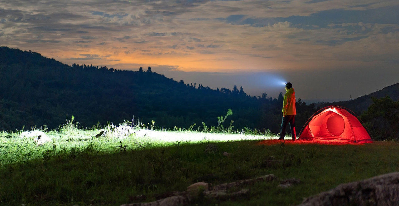 Camping de nuit avec tente rouge &eacute;clair&eacute;e et randonneur utilisant une lampe frontale puissante sur une colline au cr&eacute;puscule.