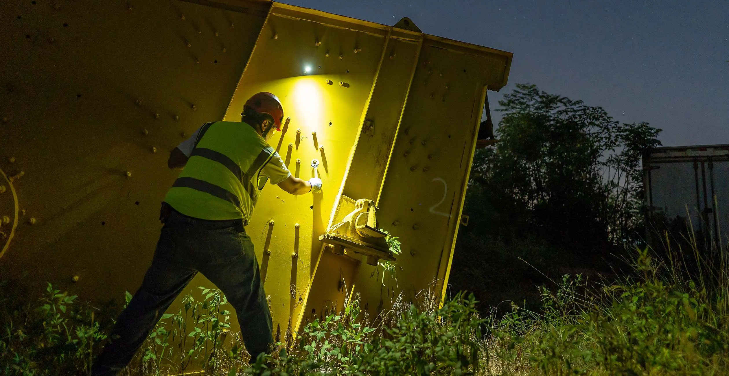 Worker wearing a hard hat and safety vest repairing heavy yellow industrial machinery with a tool at night