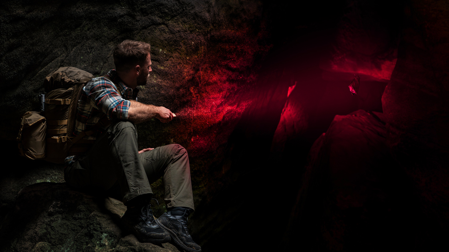 Adventurer with a tactical backpack exploring a dark cave, using a red light flashlight to observe a bat. Professional spelunking and outdoor exploration concept.