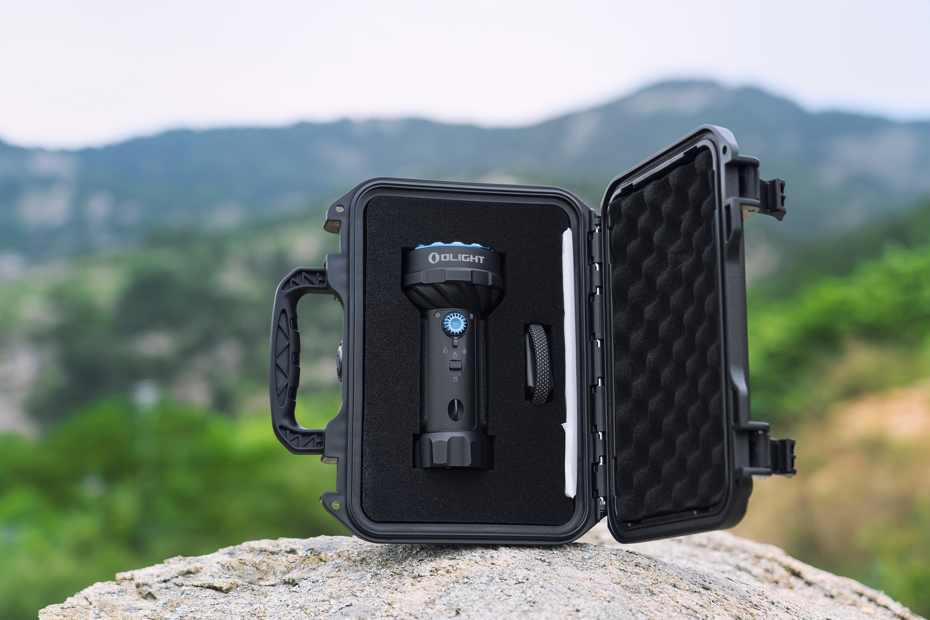 Potente linterna Olight dentro de un estuche protector abierto, posada en una roca con un paisaje montañoso y verde.