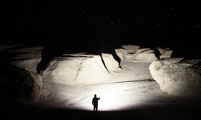 Silhouette d'un homme &eacute;clairant un paysage rocheux d&eacute;sertique la nuit avec une lampe torche puissante sous un ciel &eacute;toil&eacute;