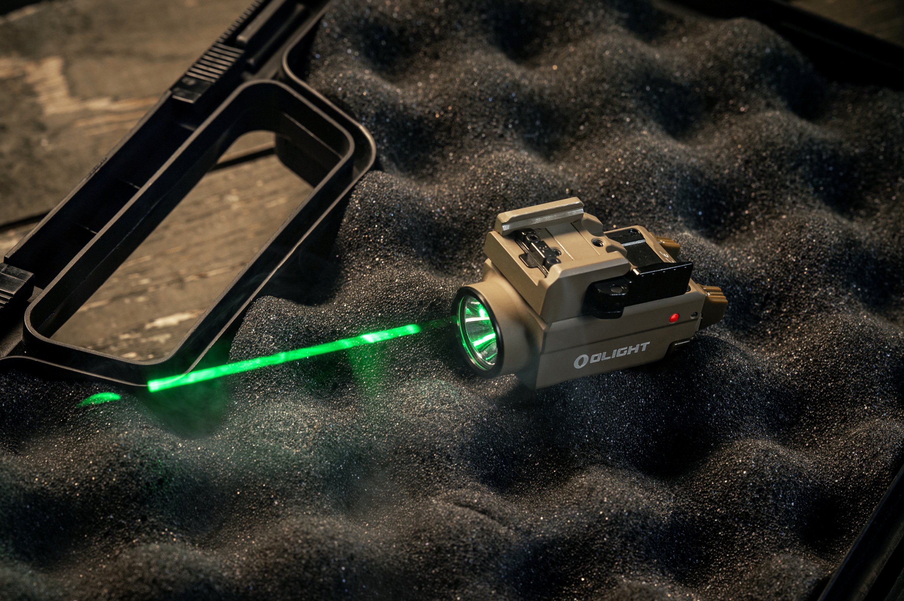 Lampe tactique Olight couleur sable avec laser vert actif, posée sur une mousse de protection noire dans un étui pour arme.