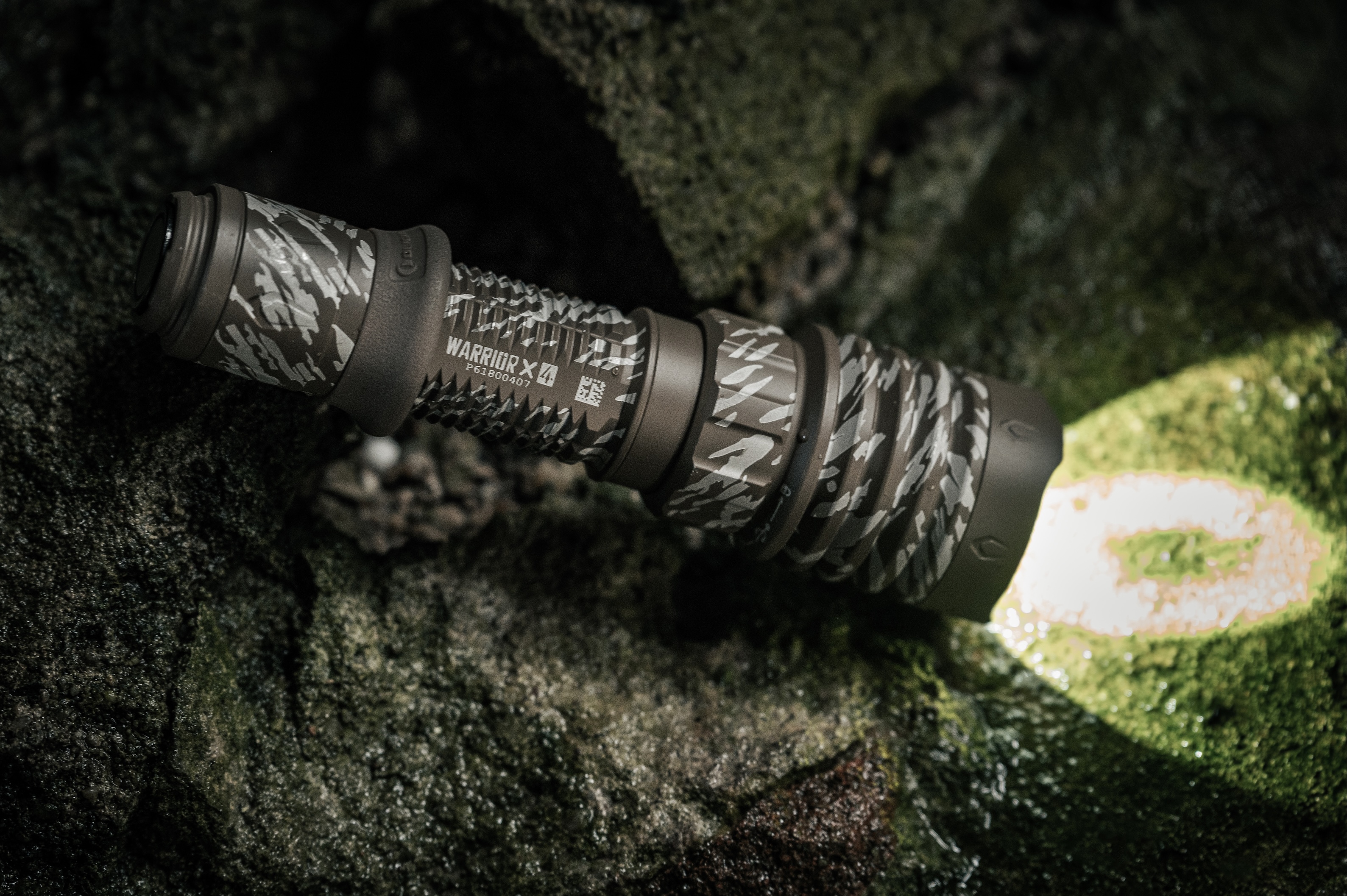 Linterna táctica Olight Warrior X 4 camuflada, iluminando un entorno rocoso y musgoso para aventuras.
