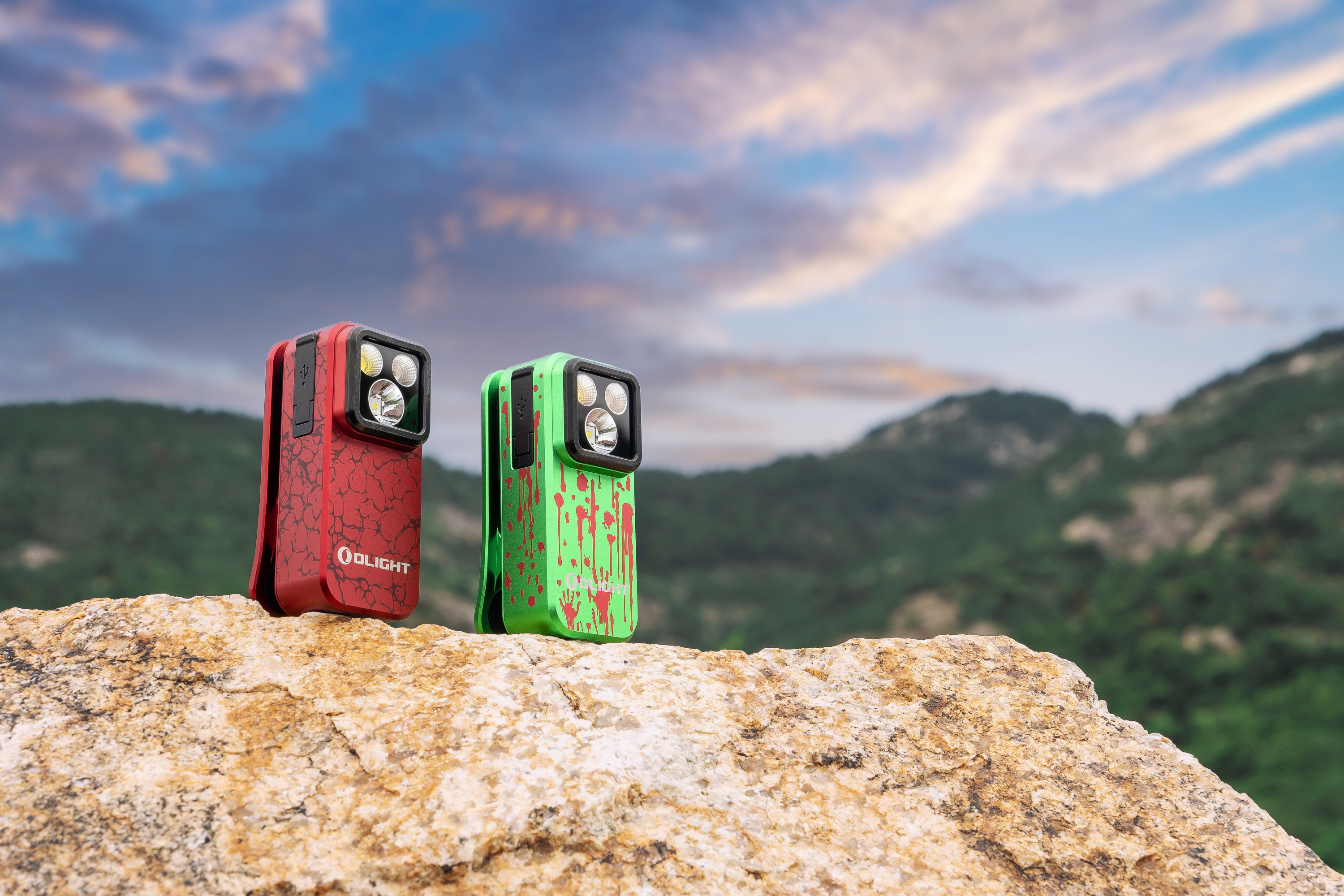 Linternas Olight rojas y verdes con patrones únicos (grietas y salpicaduras de sangre) sobre una roca en un entorno montañoso bajo un cielo nublado. Perfectas para actividades al aire libre y Halloween.