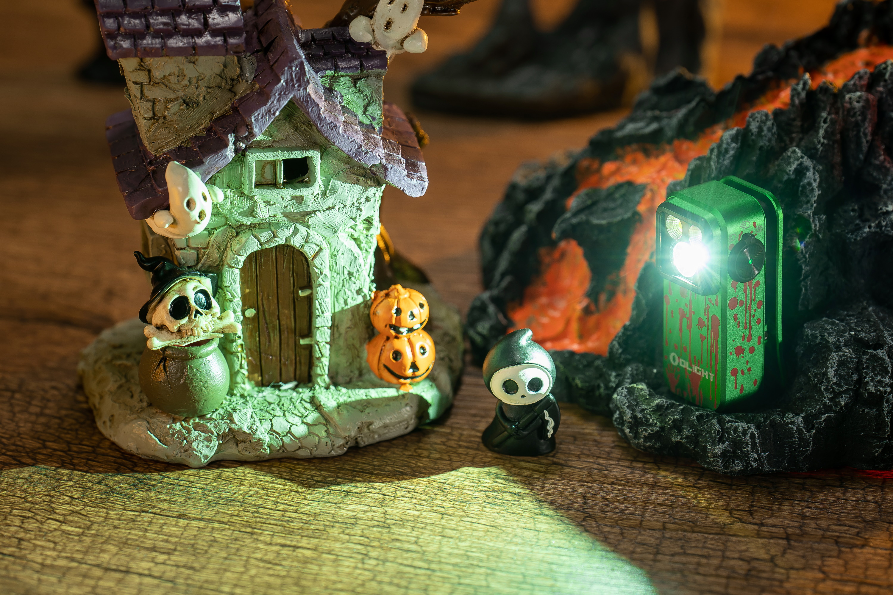 Linterna Olight verde con salpicaduras de sangre iluminando una espeluznante decoración de Halloween: casa embrujada, fantasmas, calabazas y una figura de la muerte sobre madera.