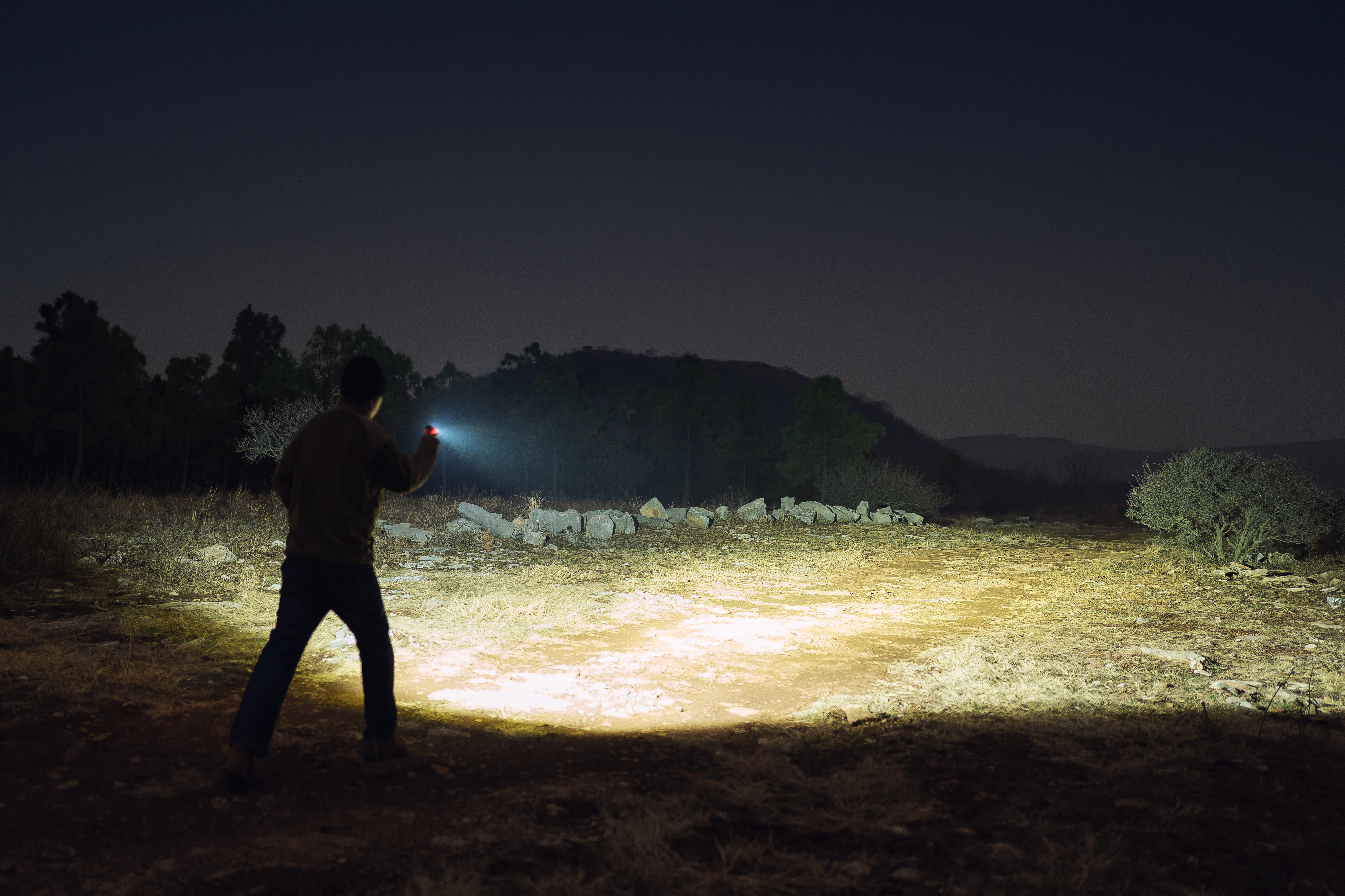 High-power portable LED flashlight used by a man at night to illuminate an outdoor field and rocky terrain.
