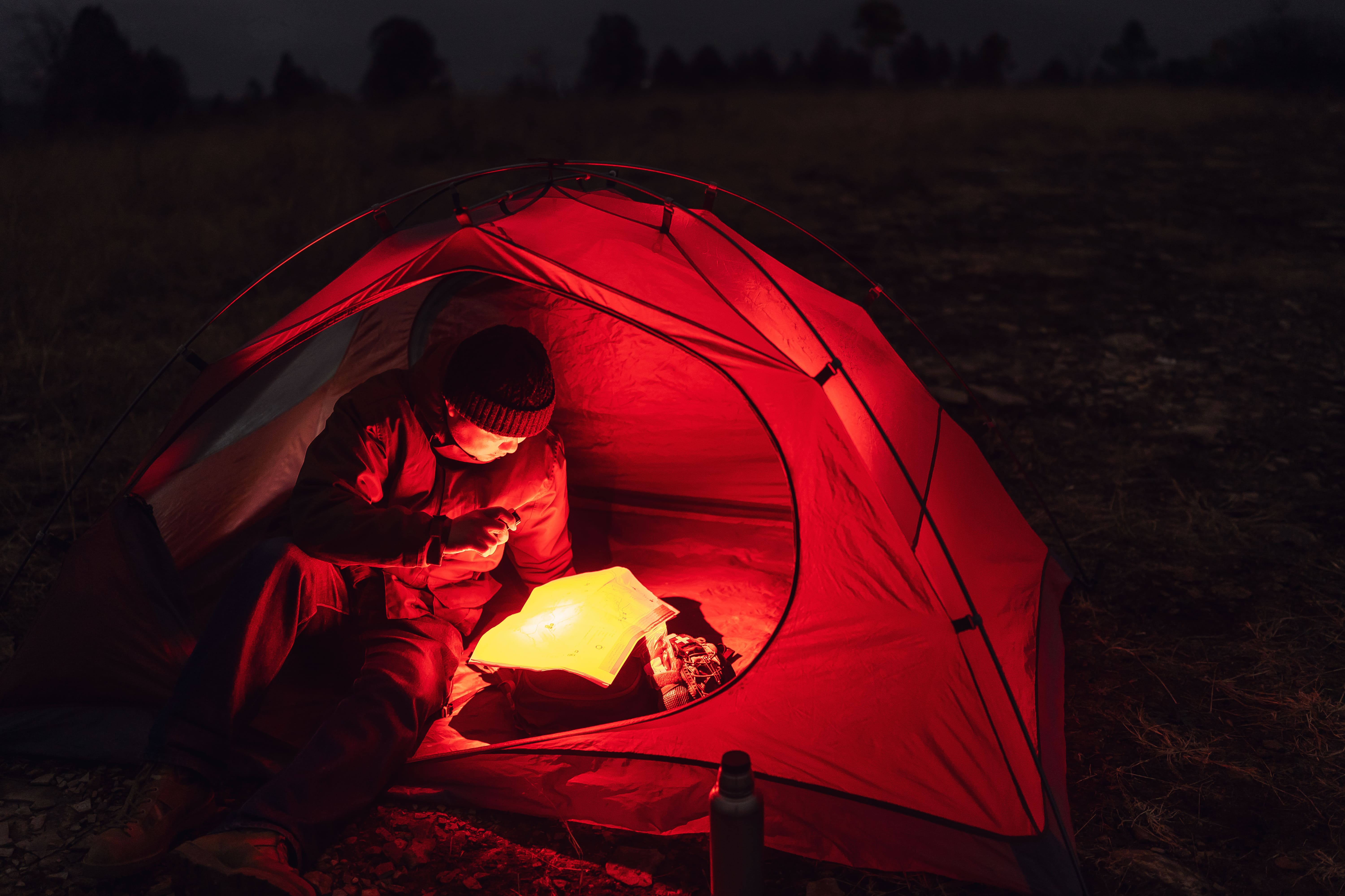 Hiker reading a map with a flashlight inside a red tent at night, illuminated by a warm red glow during a wilderness camping trip.