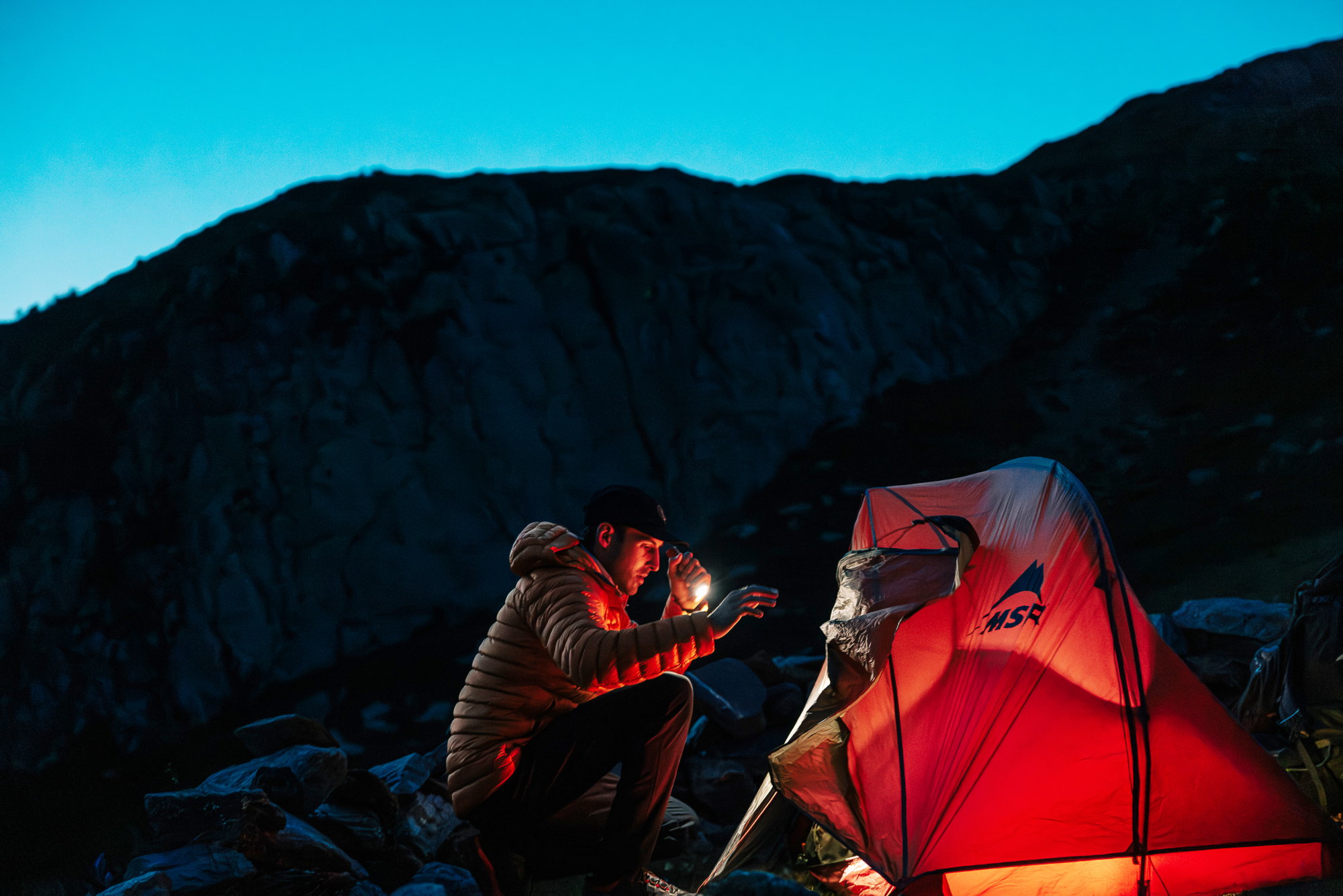 Camping de nuit en montagne avec une tente de randonnée MSR éclairée et un randonneur utilisant une lampe frontale au crépuscule.
