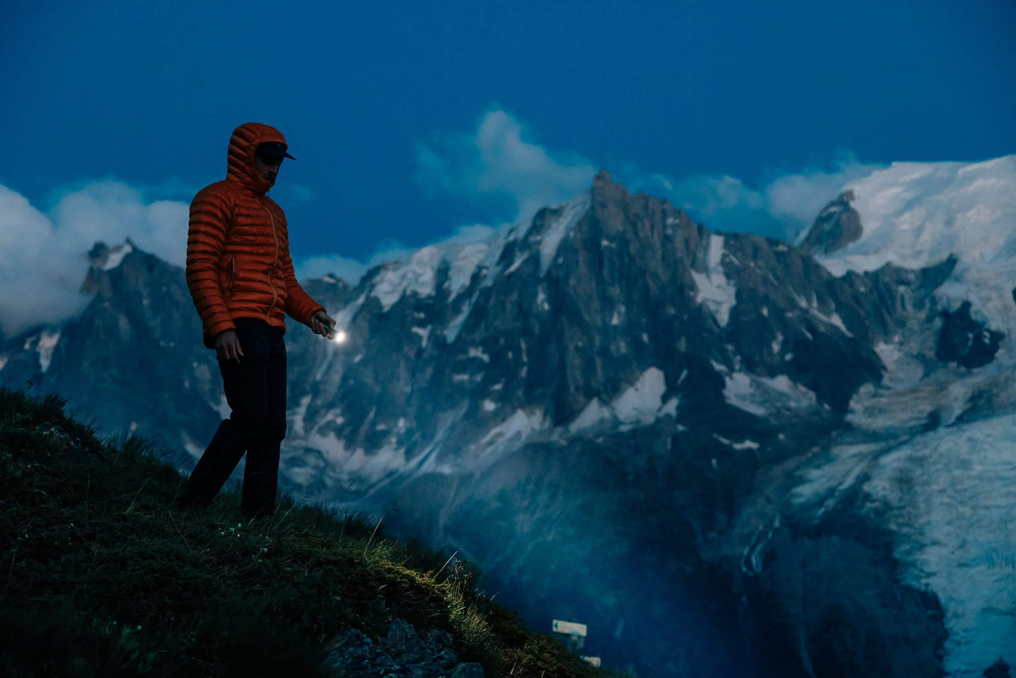 Hombre con chaqueta naranja y linterna subiendo en una ladera de montaña al atardecer, con picos nevados al fondo.