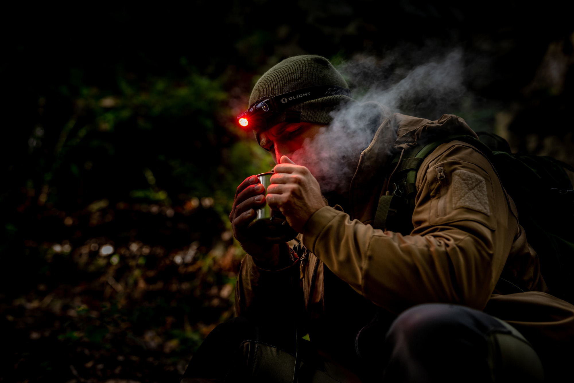 Randonneur en for&ecirc;t de nuit buvant une boisson chaude avec une lampe frontale Olight &agrave; lumi&egrave;re rouge